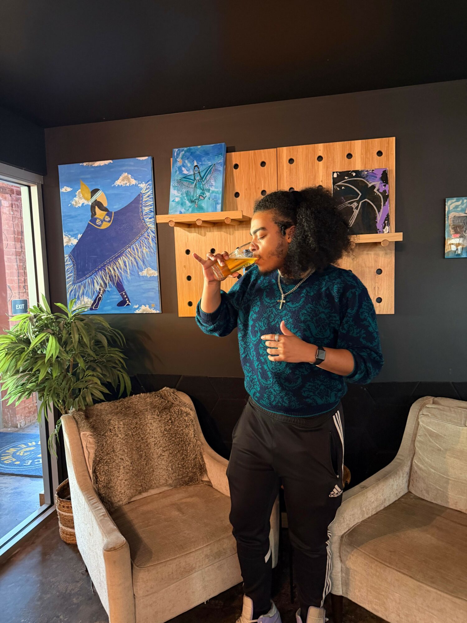 Person with curly hair wearing a blue top and black pants, standing in front of paintings, holding a phone, in a gallery.