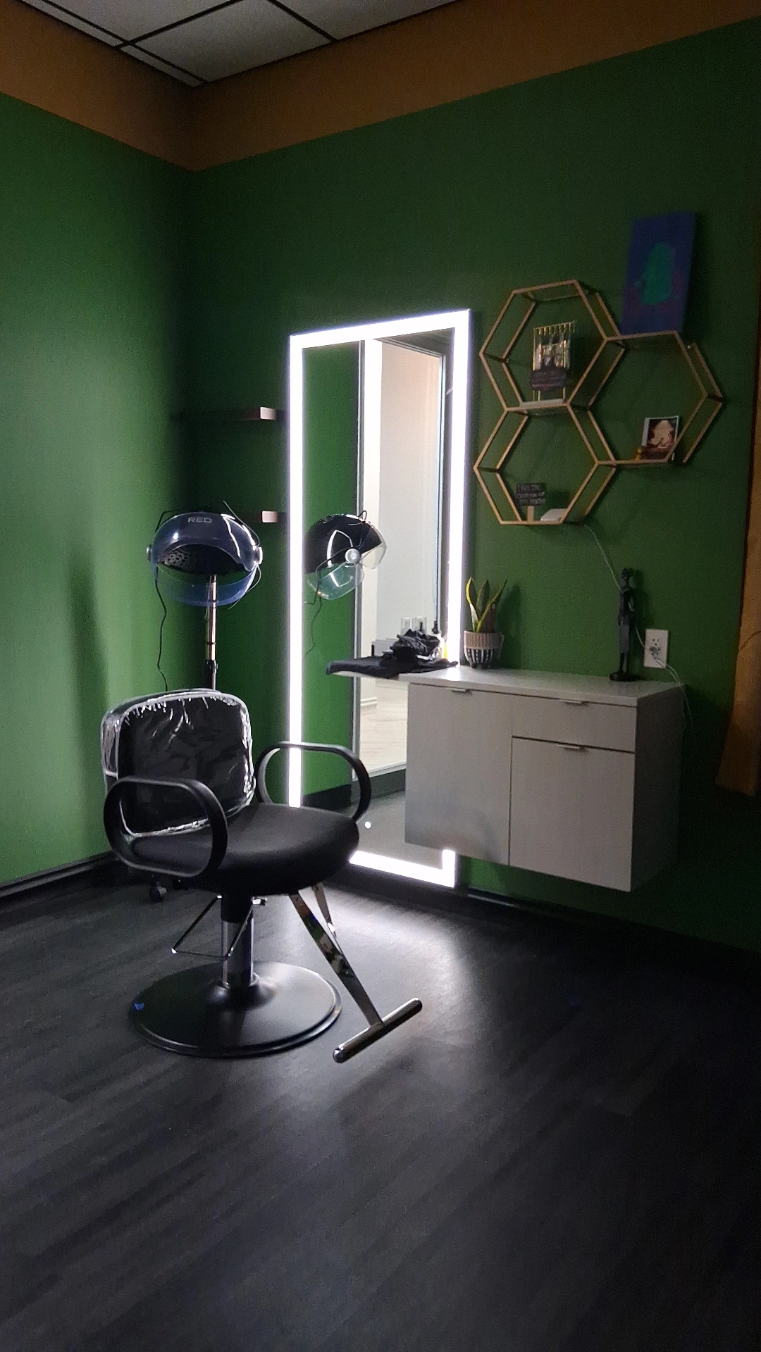 Salon with green wall, black chair, mirror, hexagonal shelves, and hair dryers, illuminated by bright light, with dark flooring.