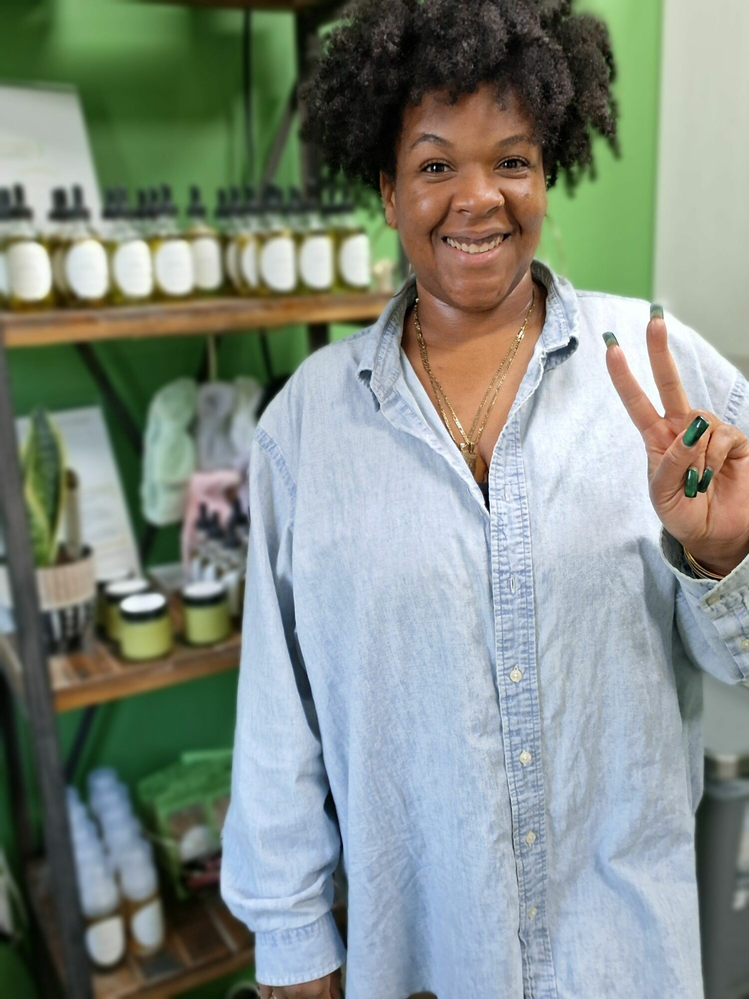 Woman with curly hair smiling and holding up a peace sign inside a store with green walls and shelves of products.