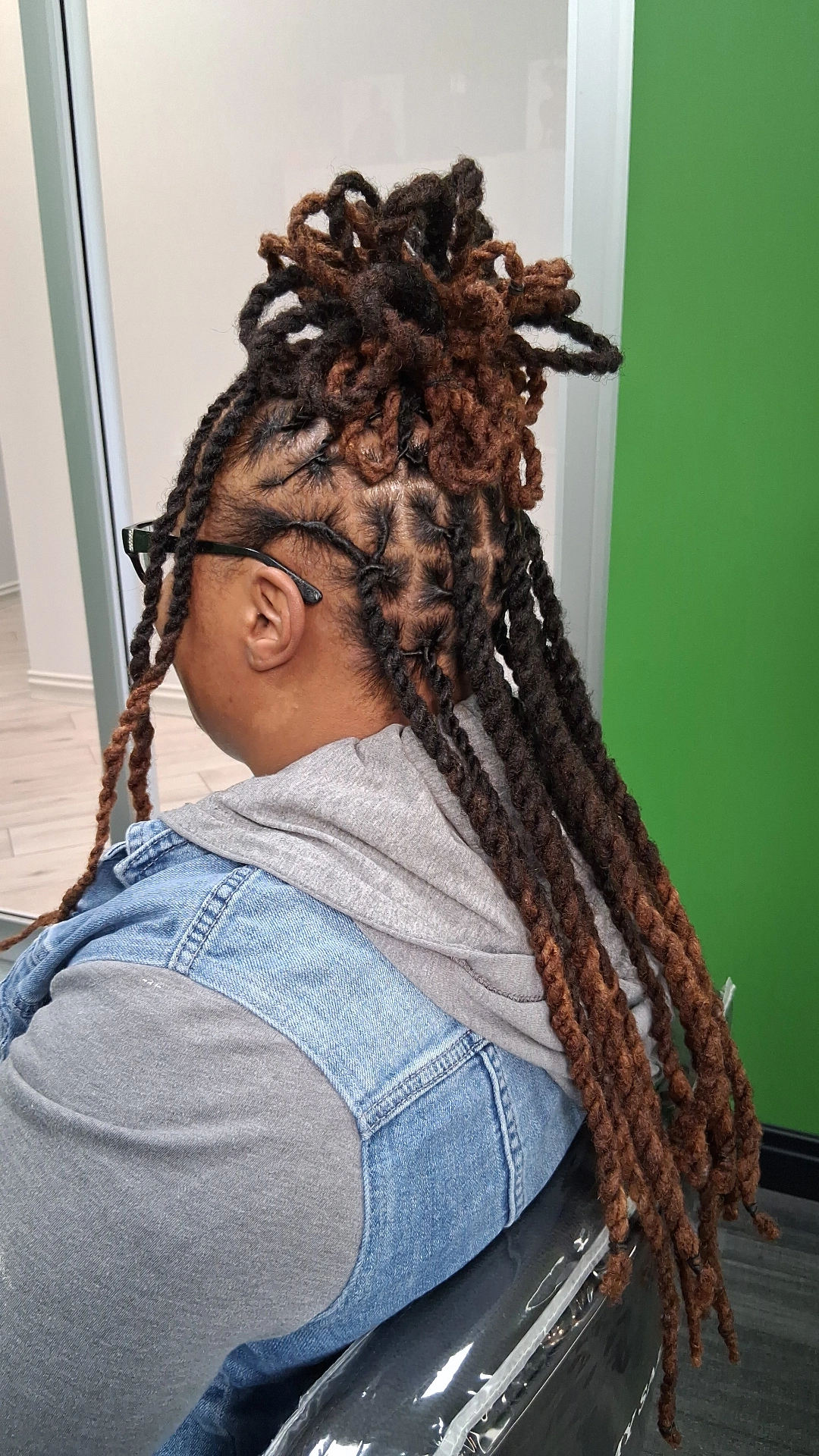 Person with styled dreadlocks sitting against a green wall, wearing a gray hoodie and denim jacket.