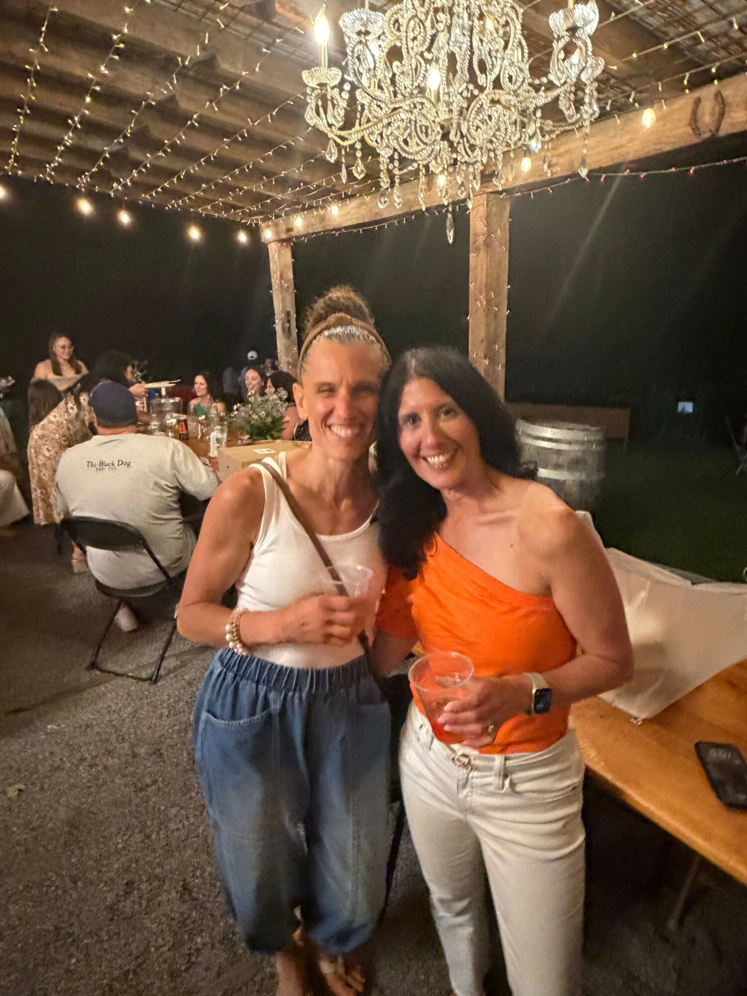 Two women standing close together, one in an orange top and white pants, the other in a white top and blue skirt, in a decorated indoor setting with a chandelier.
