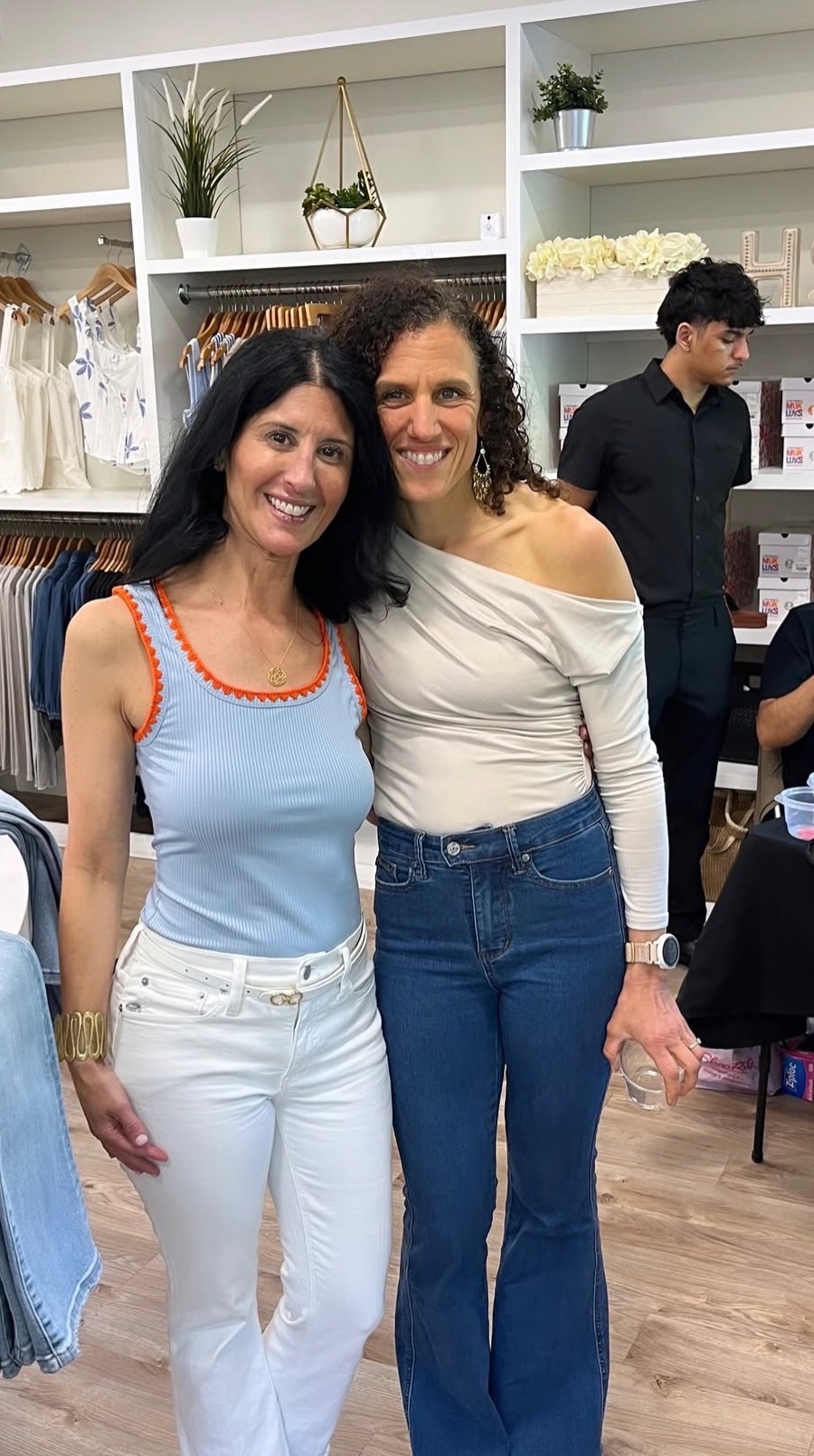 Two women smiling and standing close together in a clothing store, with shelves and other shoppers in the background.