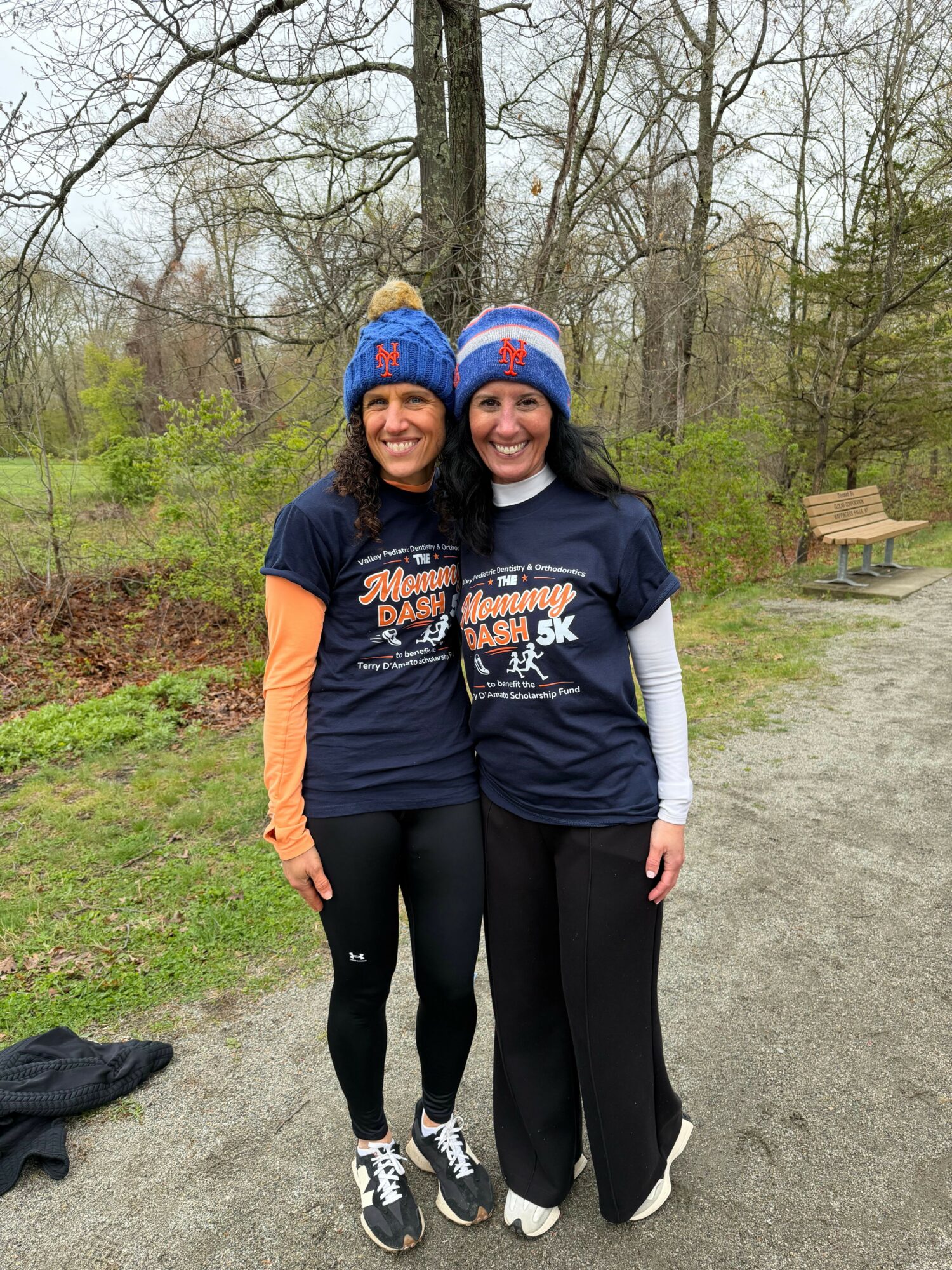 Two women standing outdoors on a path, smiling, with trees and a park bench in the background.
