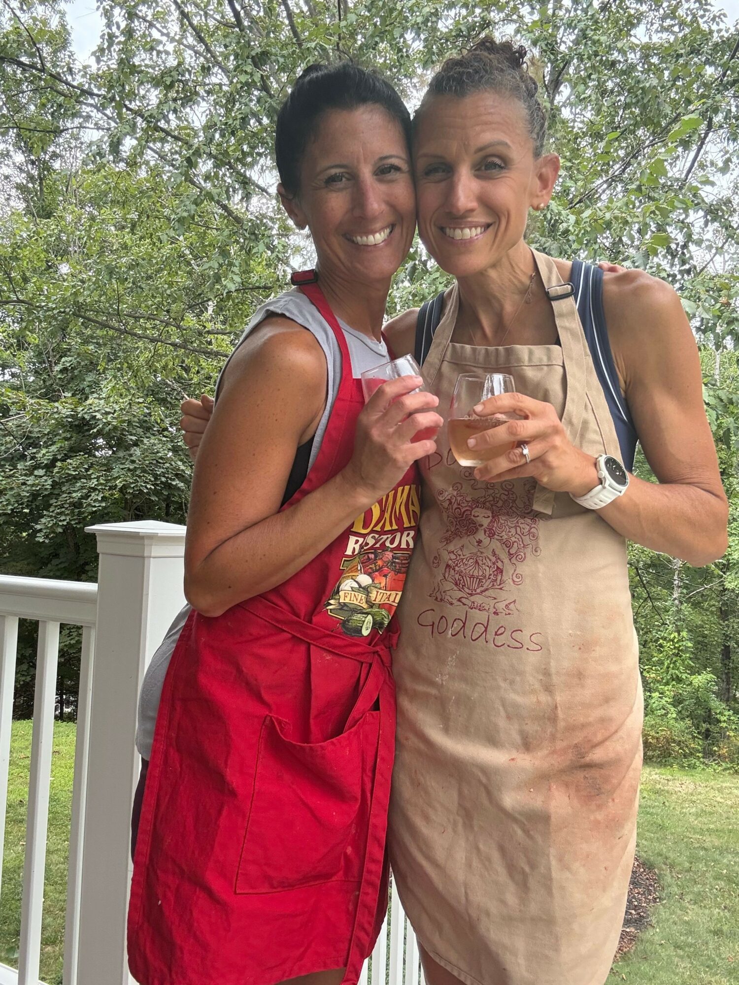 Two women standing outdoors, one wearing a beige apron and the other in a red dress, holding drinks and smiling.