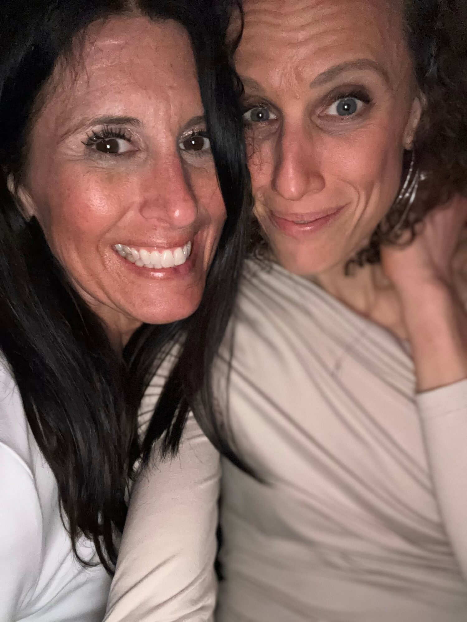Two women smiling closely, one with dark hair and the other with light hair, in a dimly lit setting.