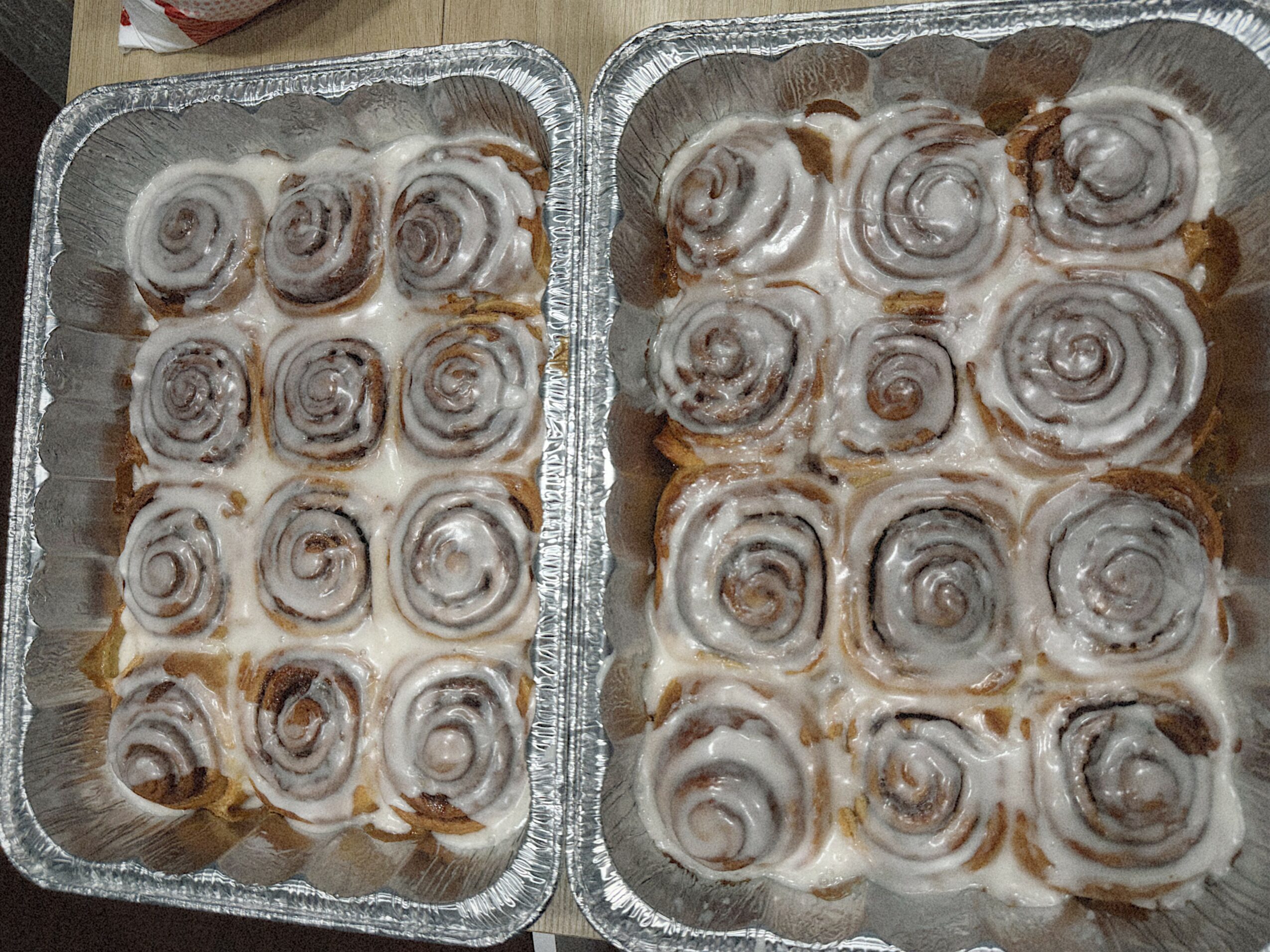 Two aluminum trays filled with cinnamon rolls topped with icing, arranged in rows, on a wooden surface.
