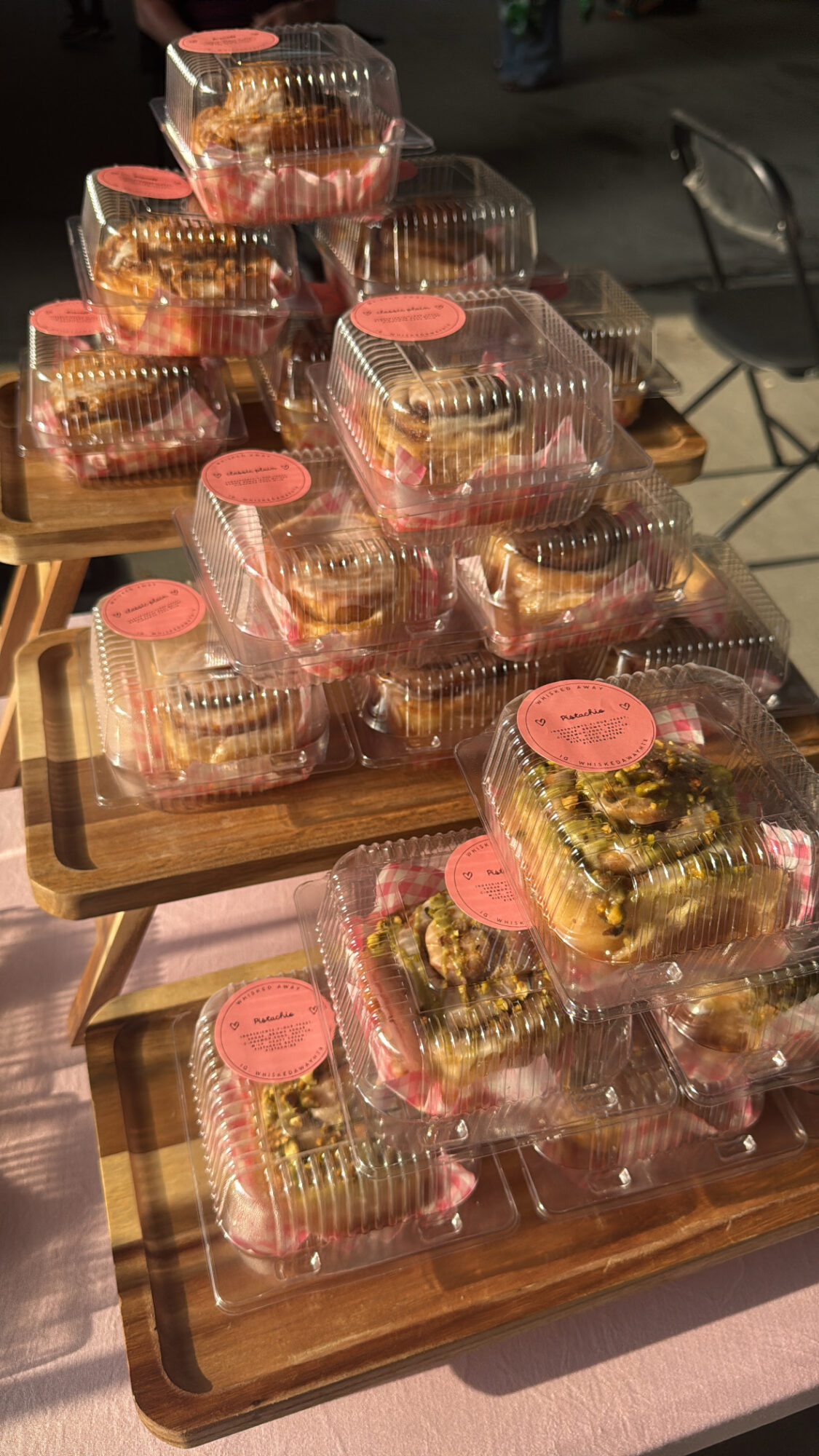 Multiple cupcakes in clear plastic containers with pink labels, arranged on wooden trays outdoors.