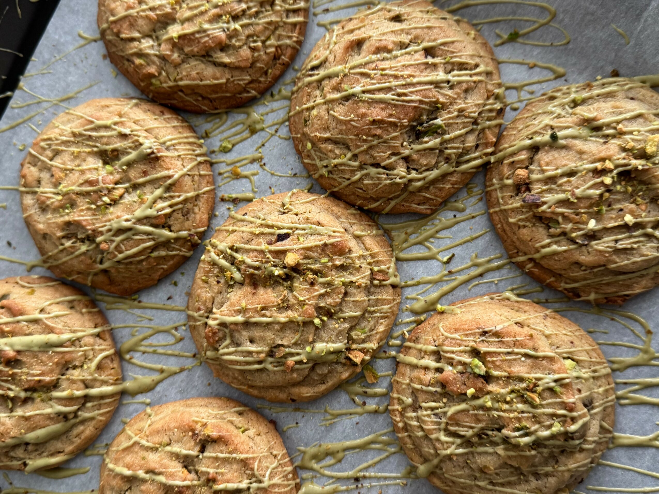 Seven round cookies with drizzle and pistachio pieces on a baking sheet, some with parchment paper, arranged in a cluster.