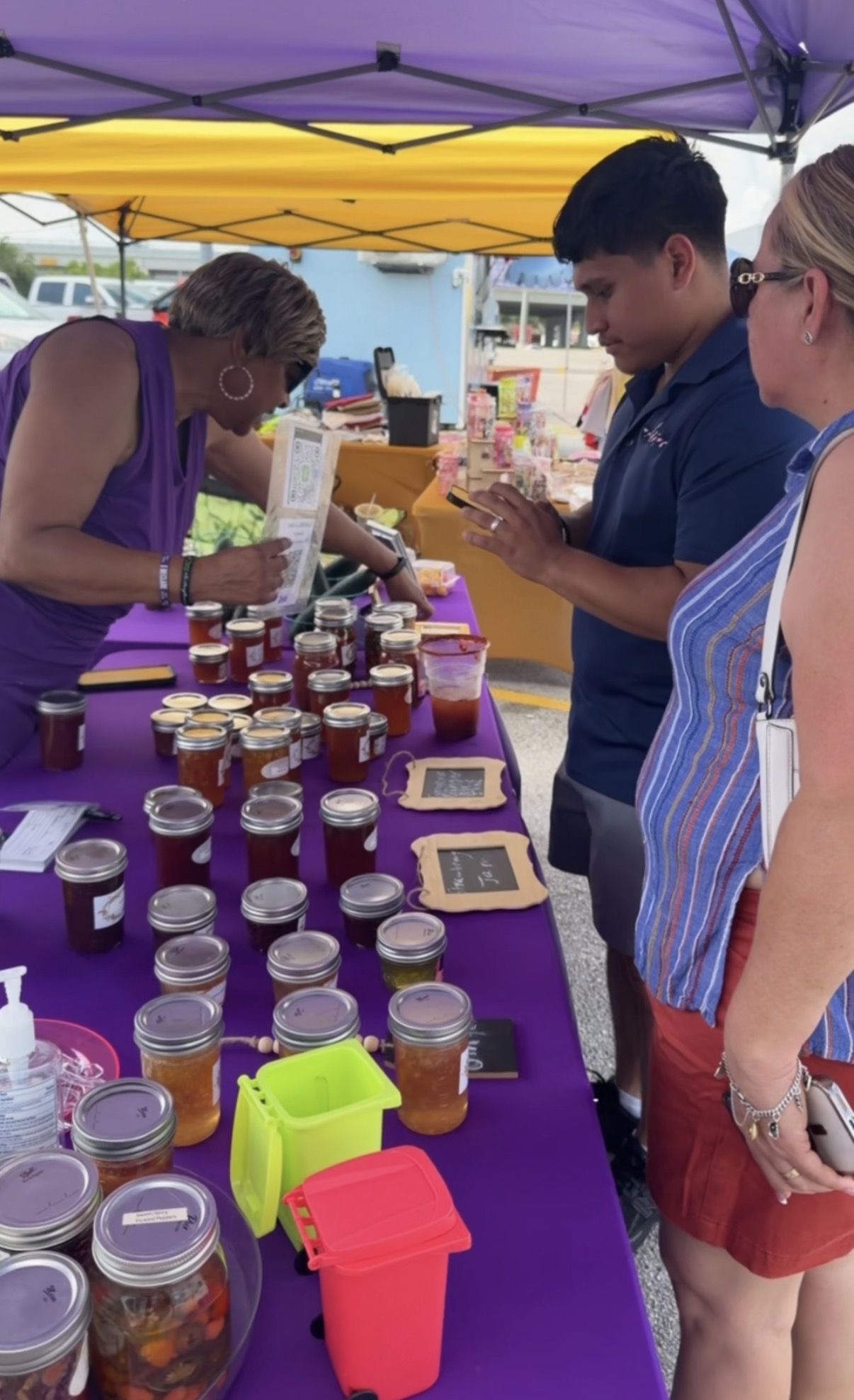 Three people at a market stall with jars of honey, under a purple and yellow canopy, engaging with products and a vendor.
