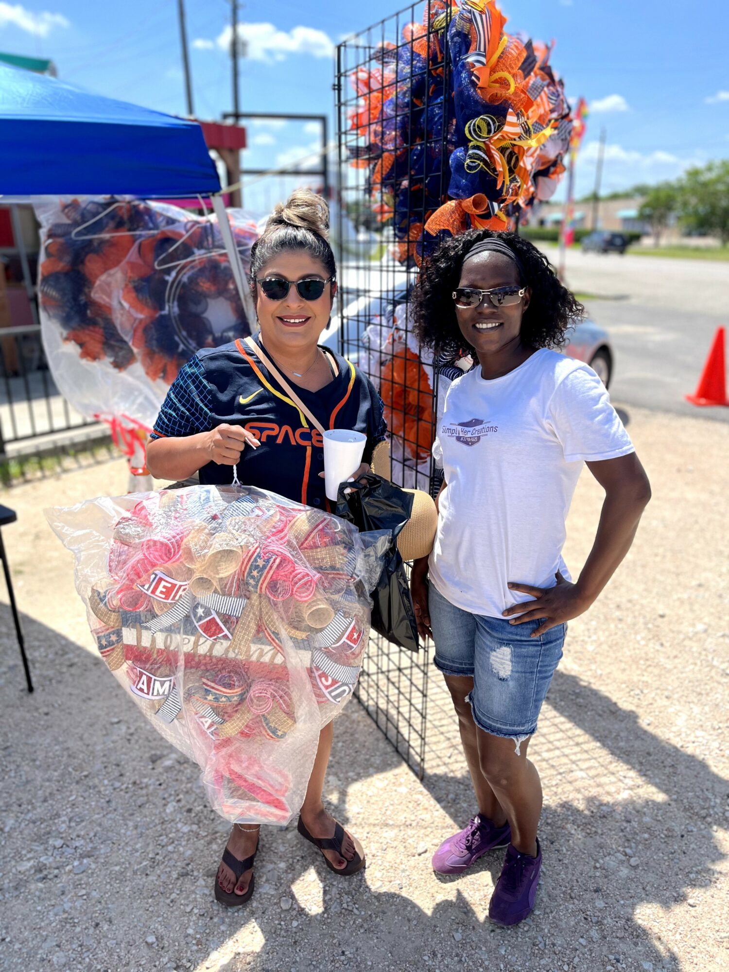 Two women standing outdoors near a vendor stall with colorful items, smiling, one holding a large bag, sunny day.