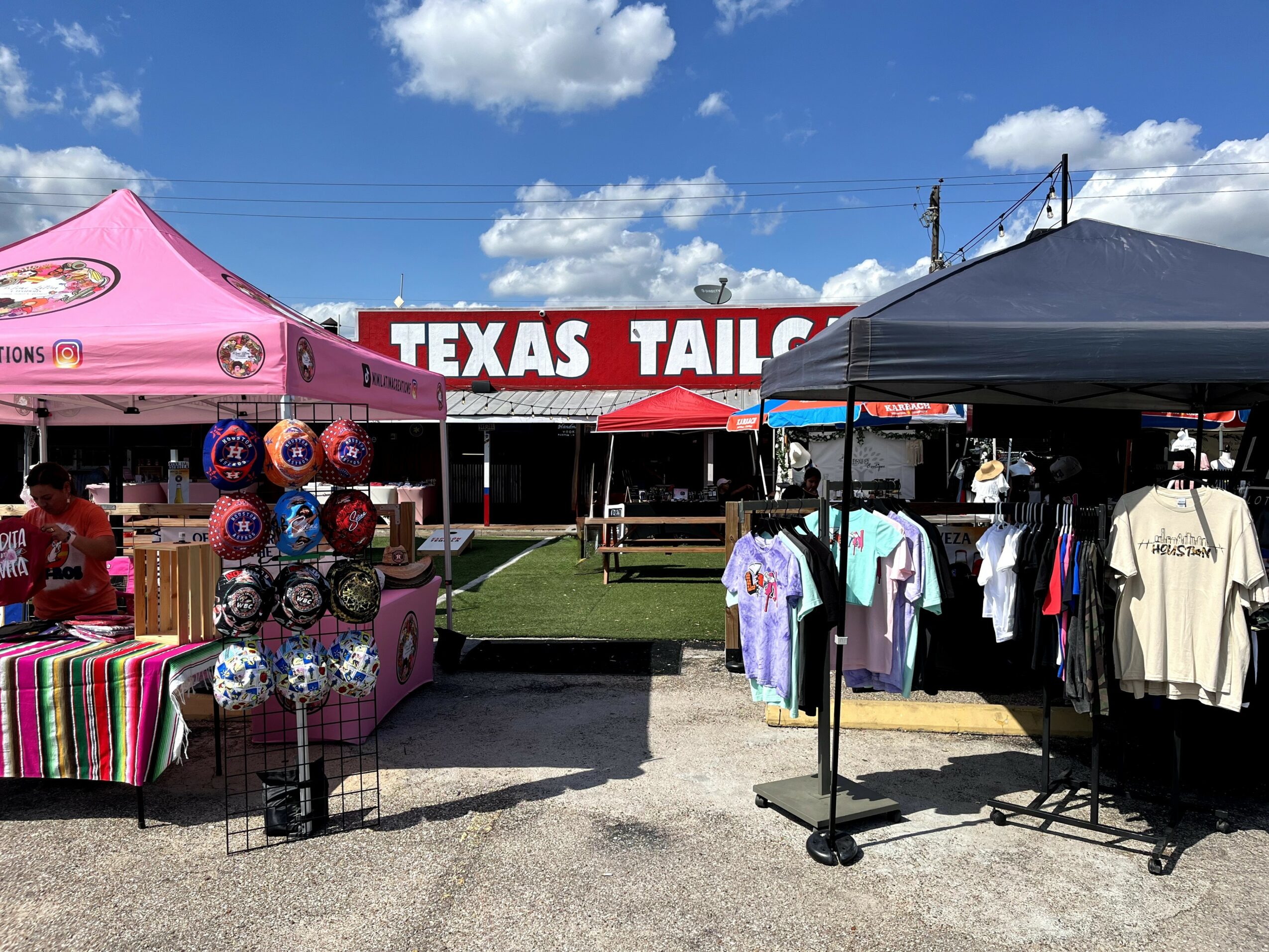 Outdoor market with tents, clothing, and hats for sale, under a partly cloudy sky, with a Texas Tailgate sign in the background.