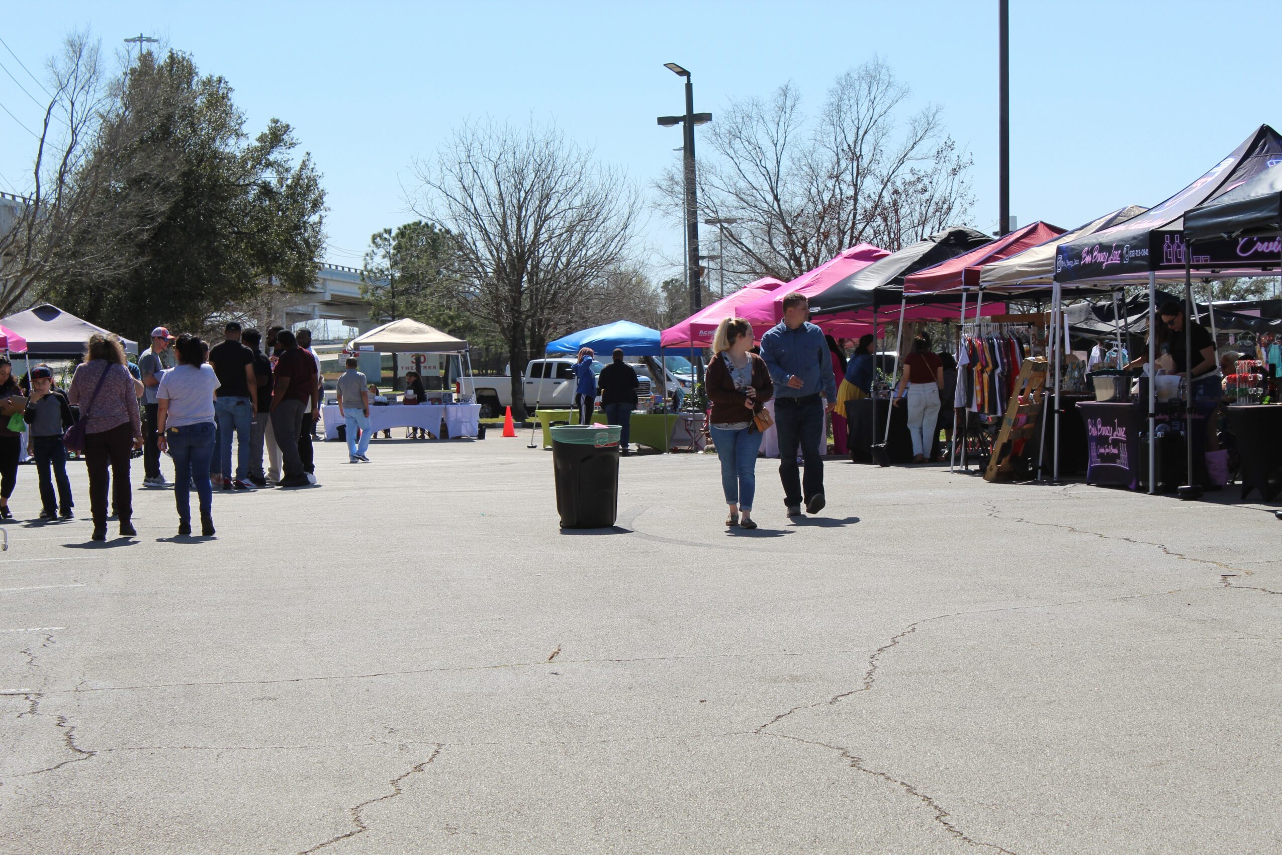 Outdoor market with tents and people browsing, trees and a clear sky in the background.