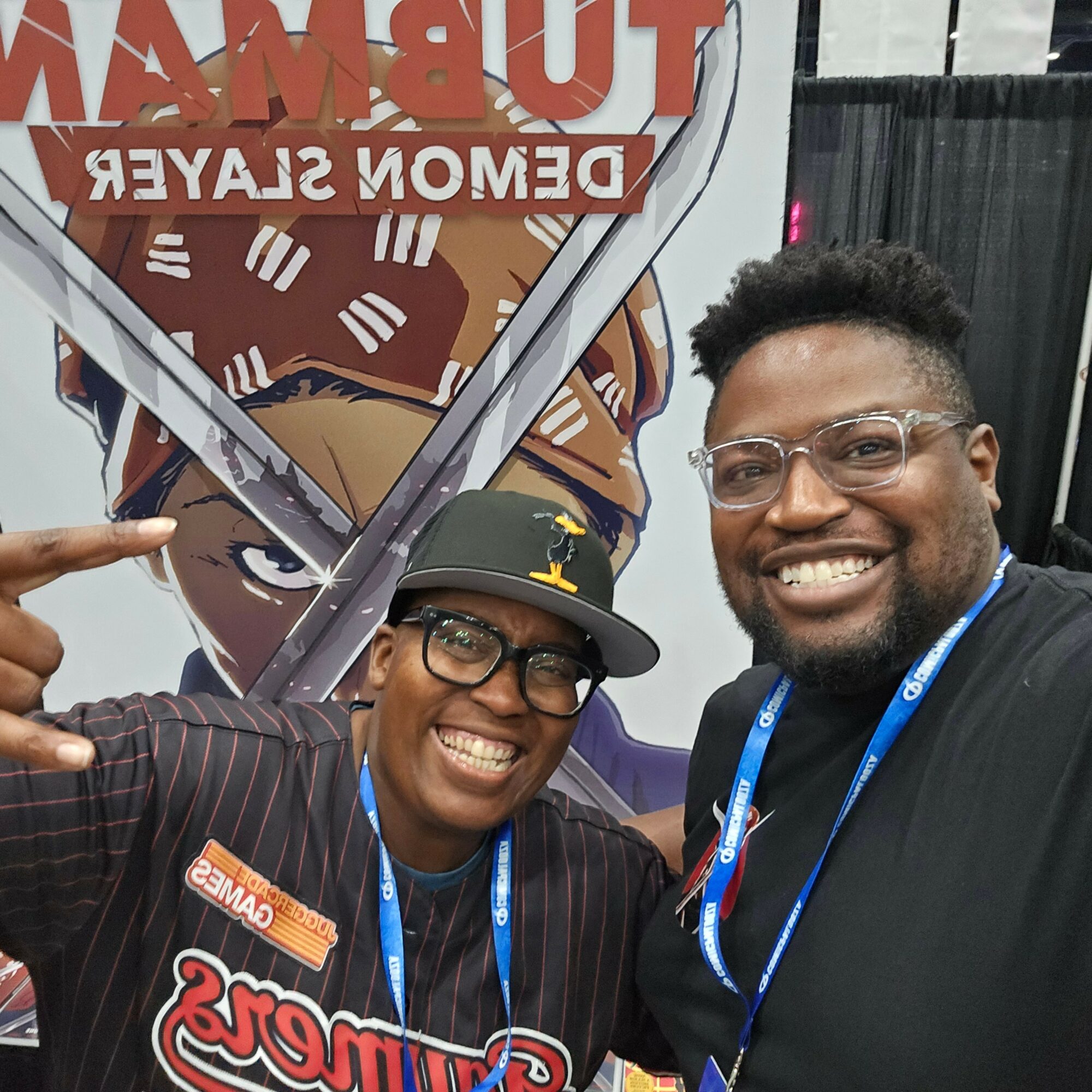 Two smiling people taking a selfie in front of a large anime-themed banner, one wearing glasses and a cap, the other with glasses and a black shirt.