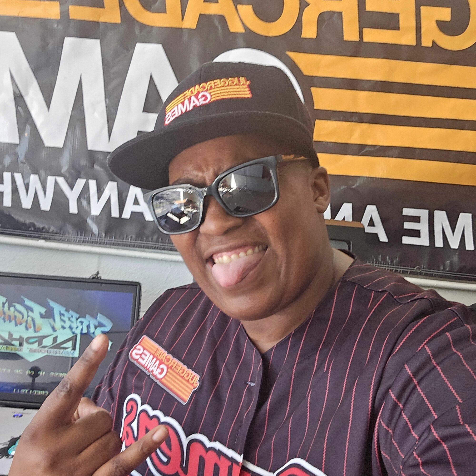 Person wearing sunglasses and a cap, smiling and making a hand gesture, in front of a sign with yellow and black stripes.