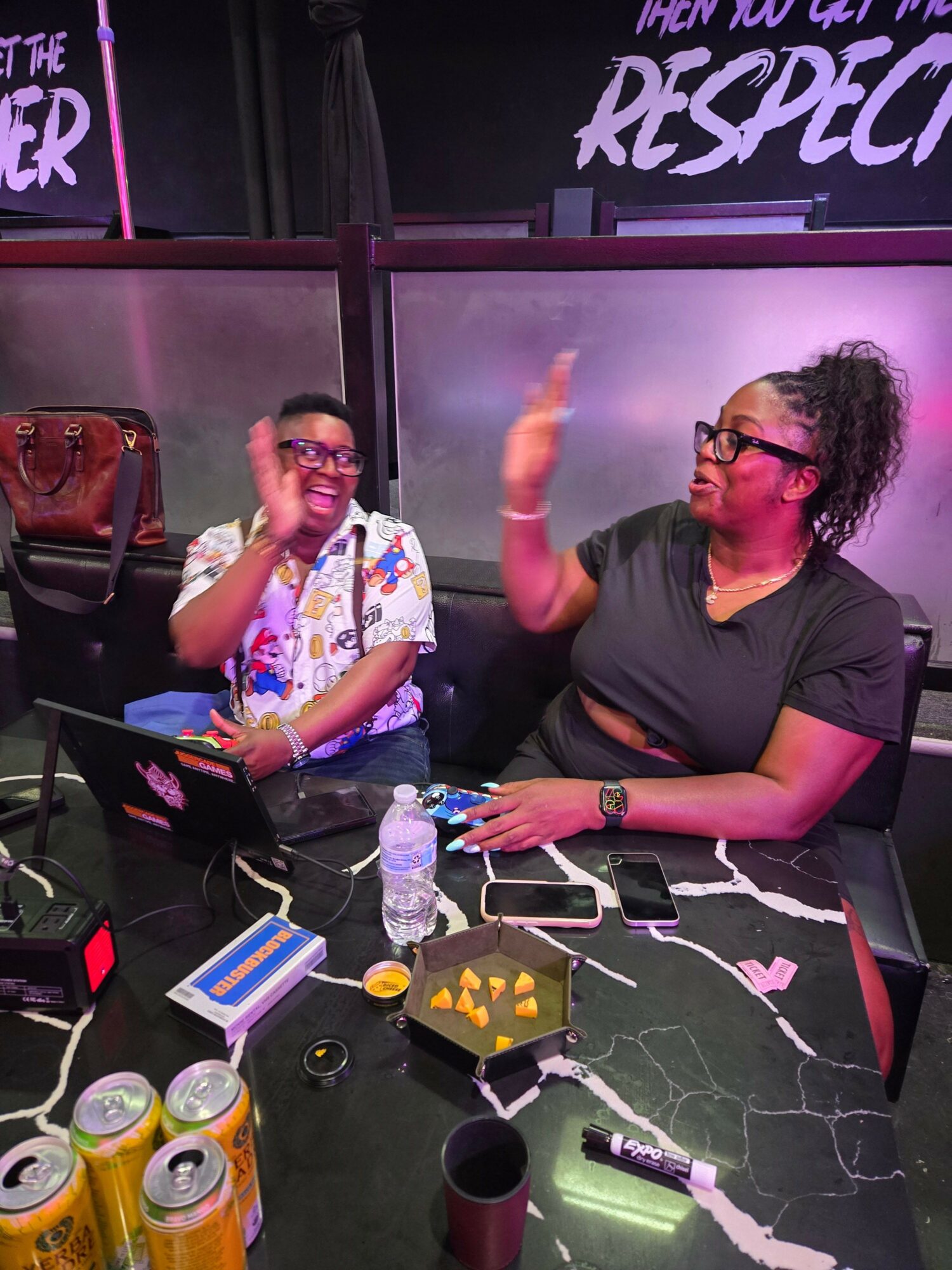 Two women sit at a table with drinks and snacks, one woman gestures while the other listens, in a room with purple lighting.