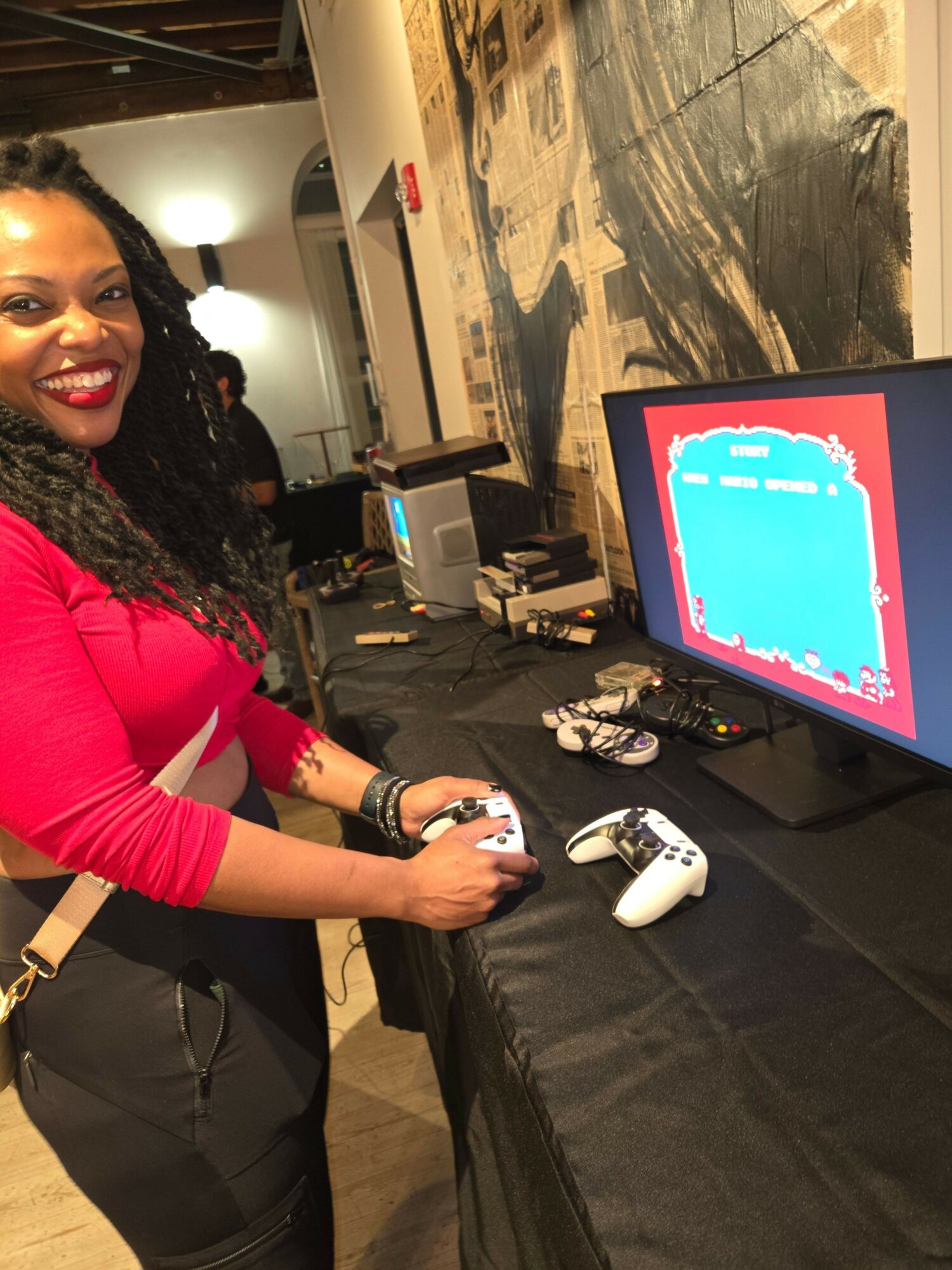 Person with long curly hair wearing a red top playing a video game at a table with a monitor and gaming controllers.