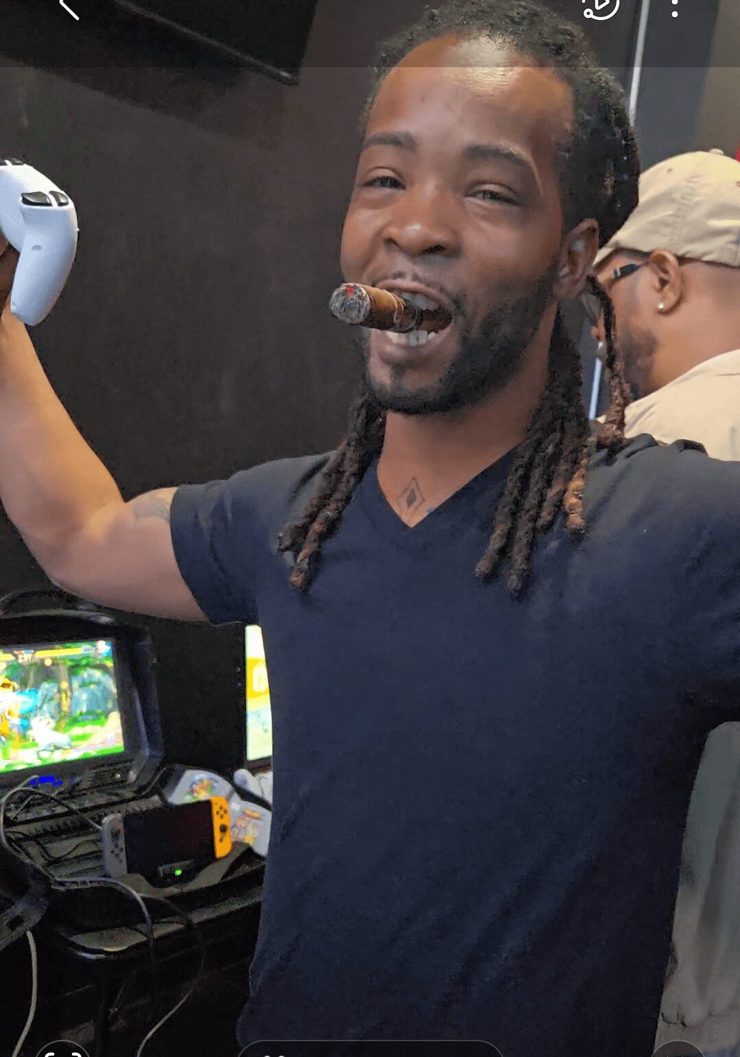 Man with dreadlocks smiling, holding a gaming controller, in a room with gaming equipment and another person in the background.
