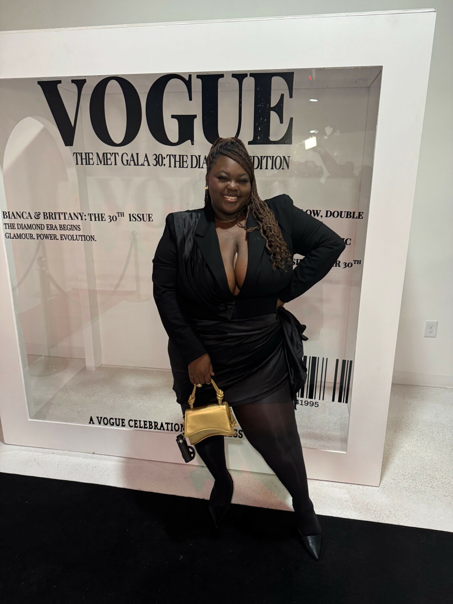 Woman in black outfit holding a yellow bag, standing in front of a Vogue display, facing sideways.