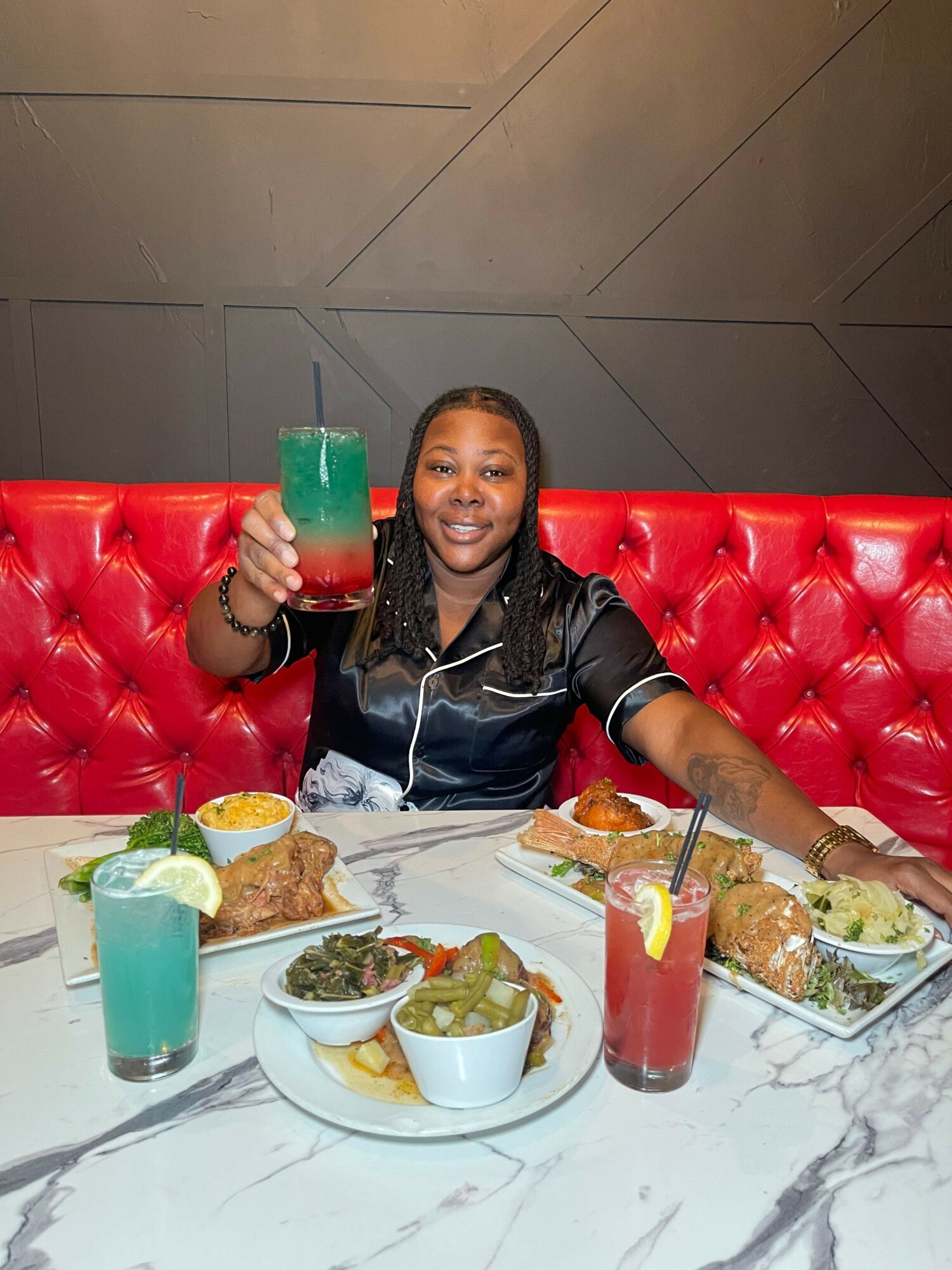 Person sitting at a table with food and drinks, holding a red drink, smiling, on a red cushioned seat.