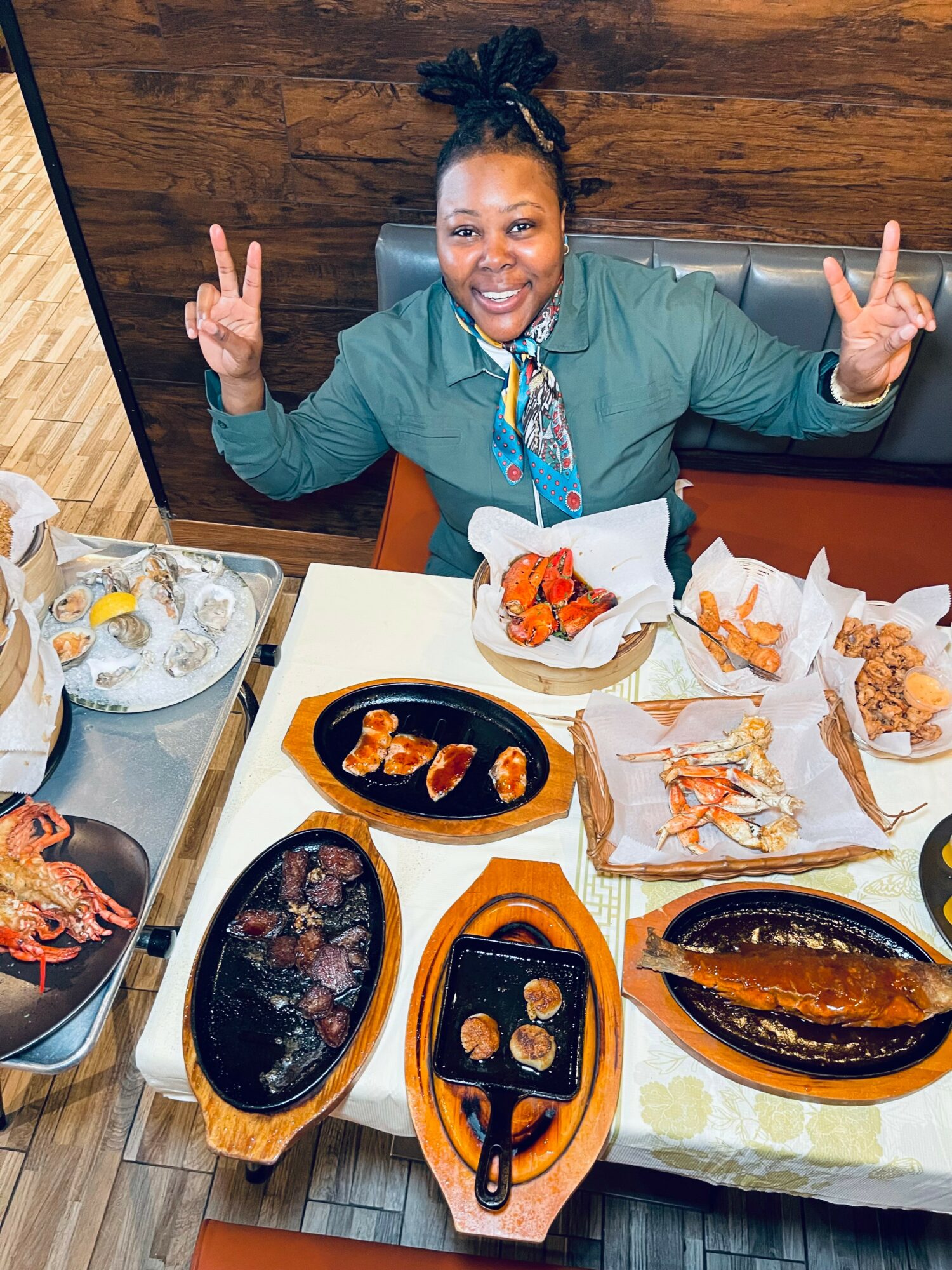 Woman smiling at a table with various dishes, making peace signs with both hands.
