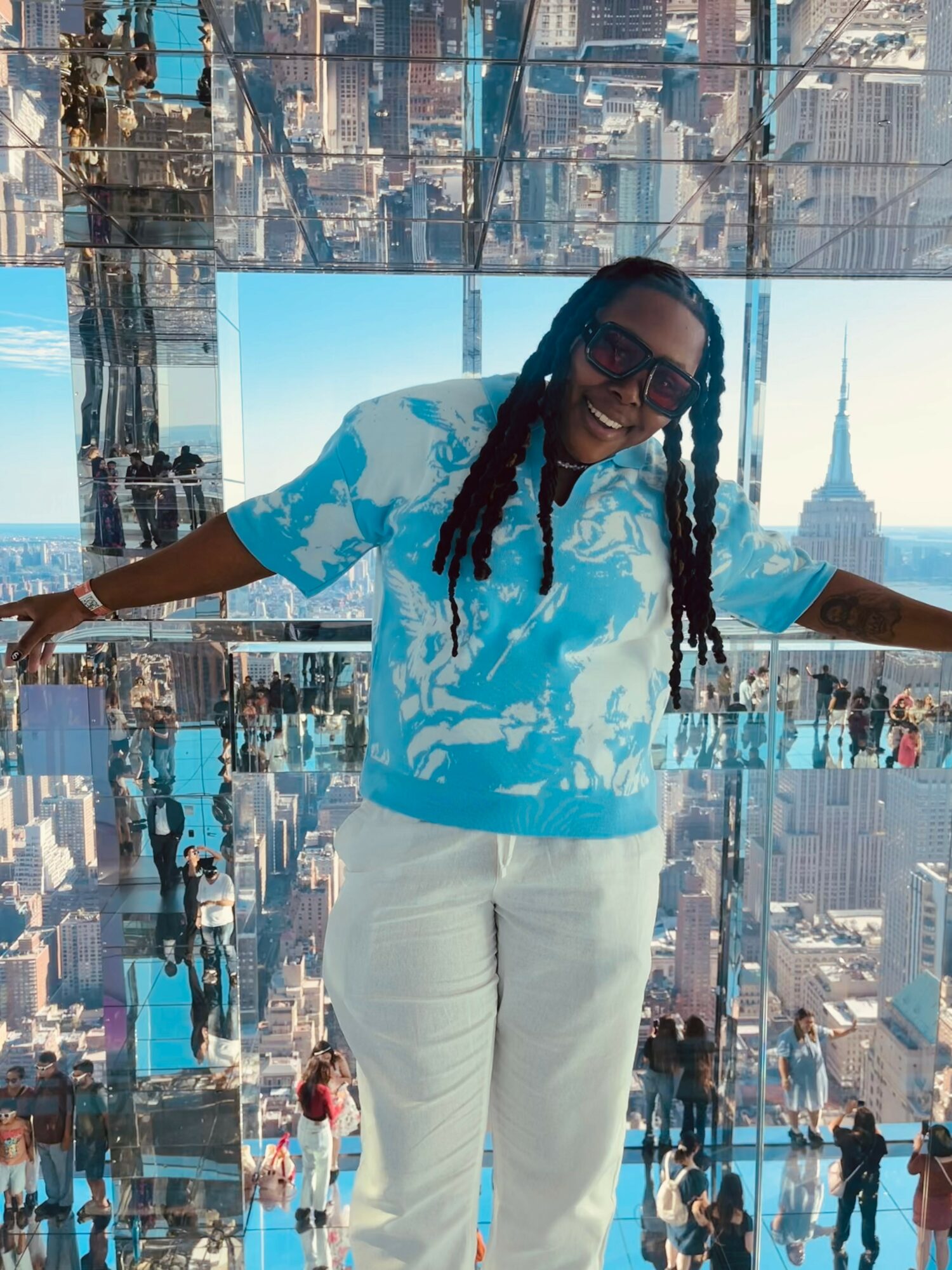 Person with long hair and sunglasses smiling, standing with arms outstretched inside a mirrored structure overlooking cityscape.