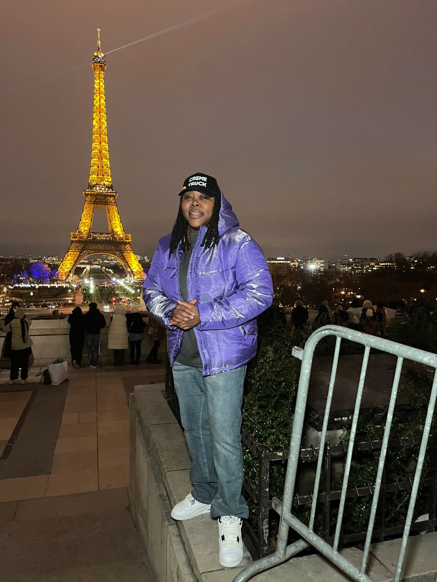 Person in purple jacket and jeans standing on balcony at night with city lights and a tall, illuminated structure in the background.