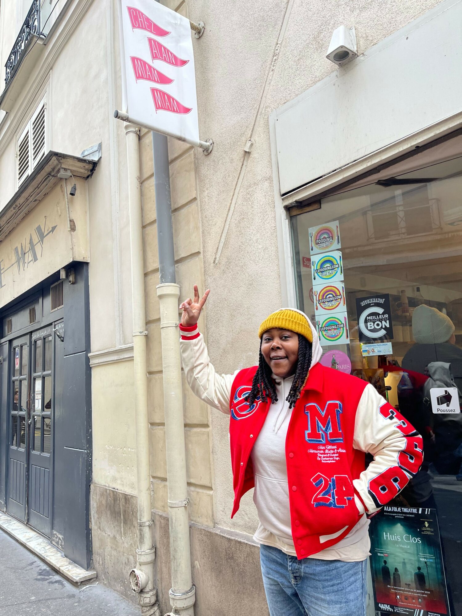Person wearing a red and white jacket and yellow beanie making a peace sign outside a building.