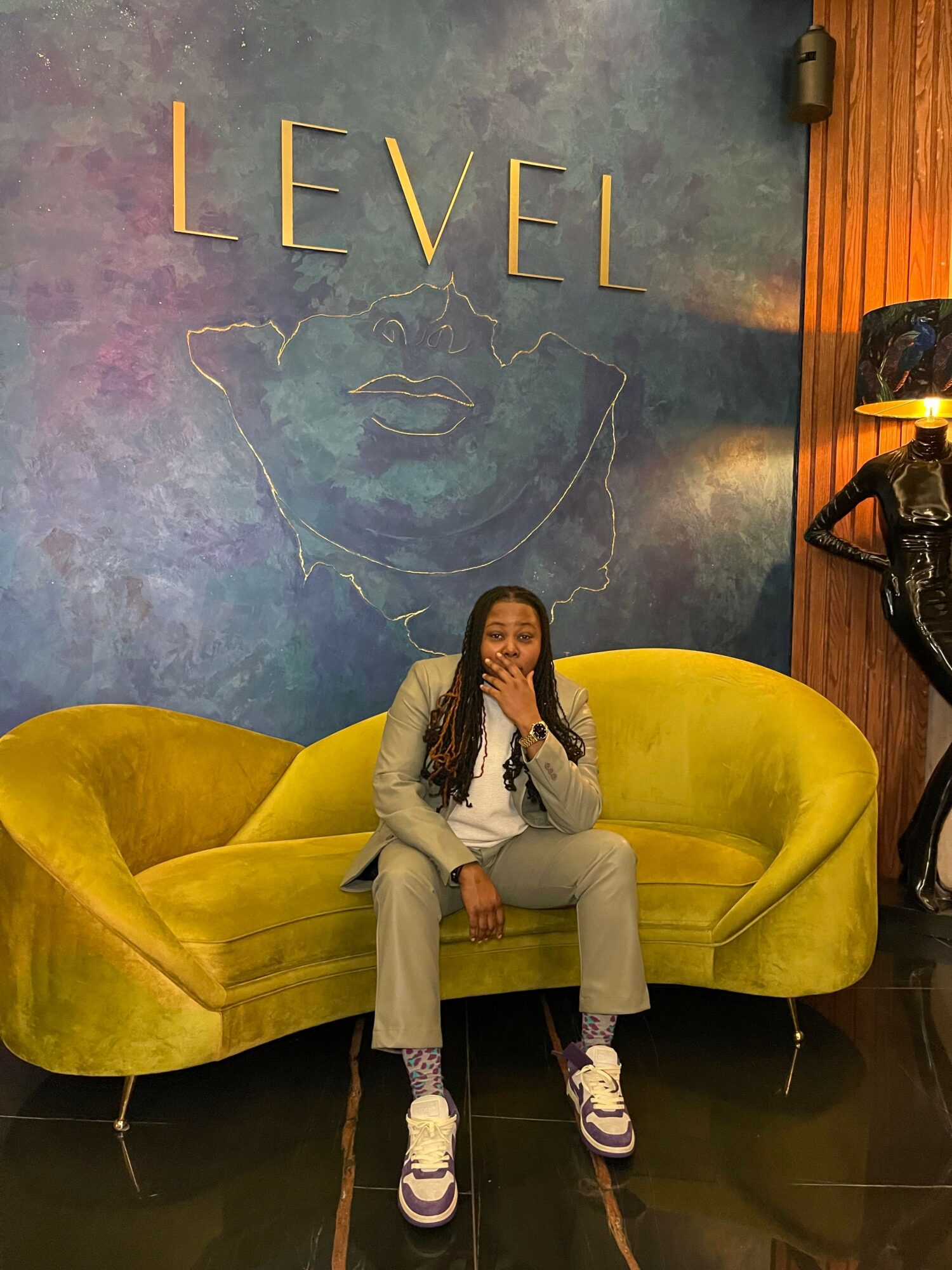 Person with long hair sitting on a yellow sofa in front of a colorful wall with the word 'LEVEL' and a face outline.