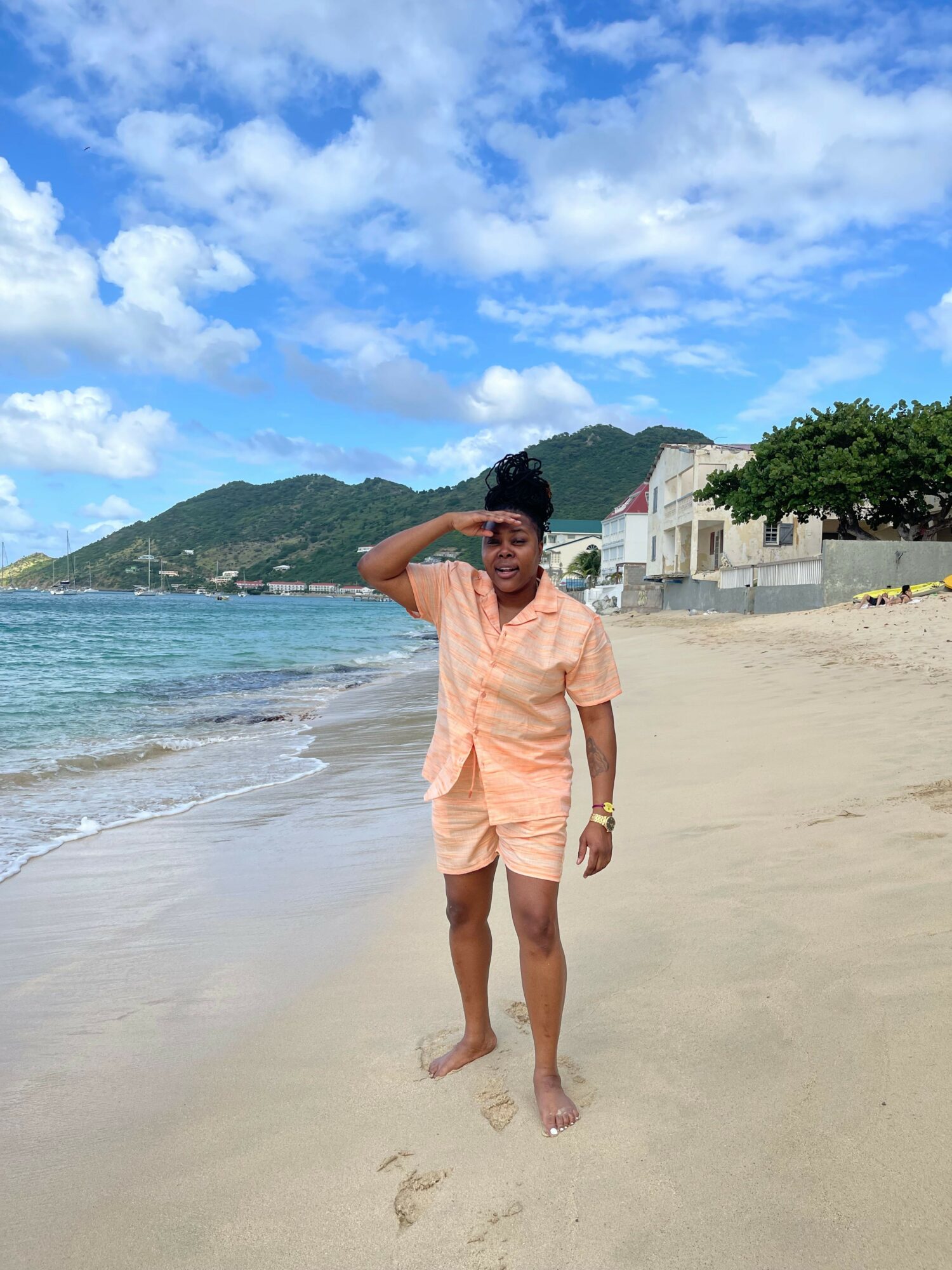 Woman in peach dress walking on sandy beach with ocean, sky, and hills in background.