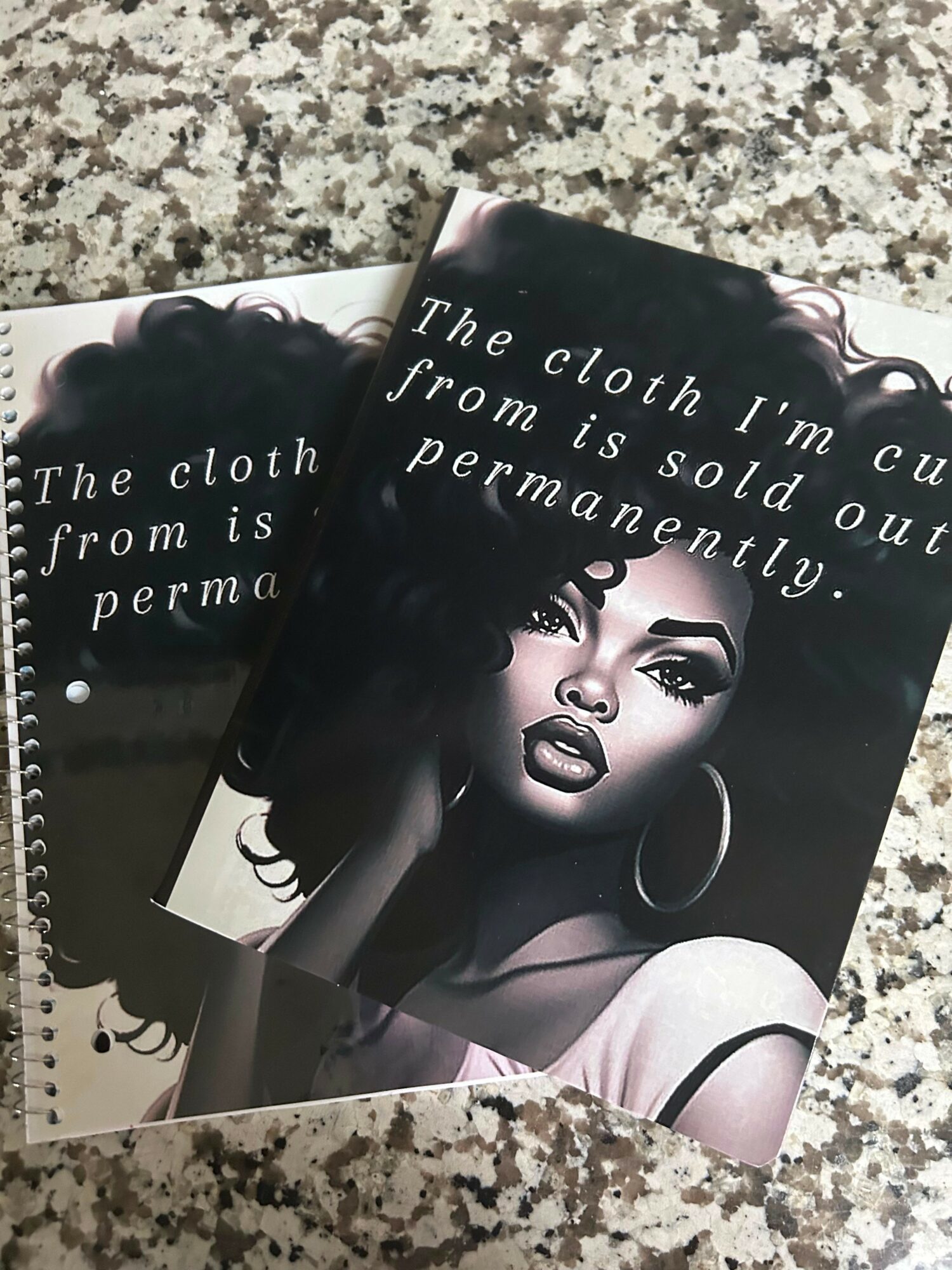 Two booklets with a black-and-white portrait of a woman with large hoop earrings, curly hair, and bold makeup, on a granite surface.