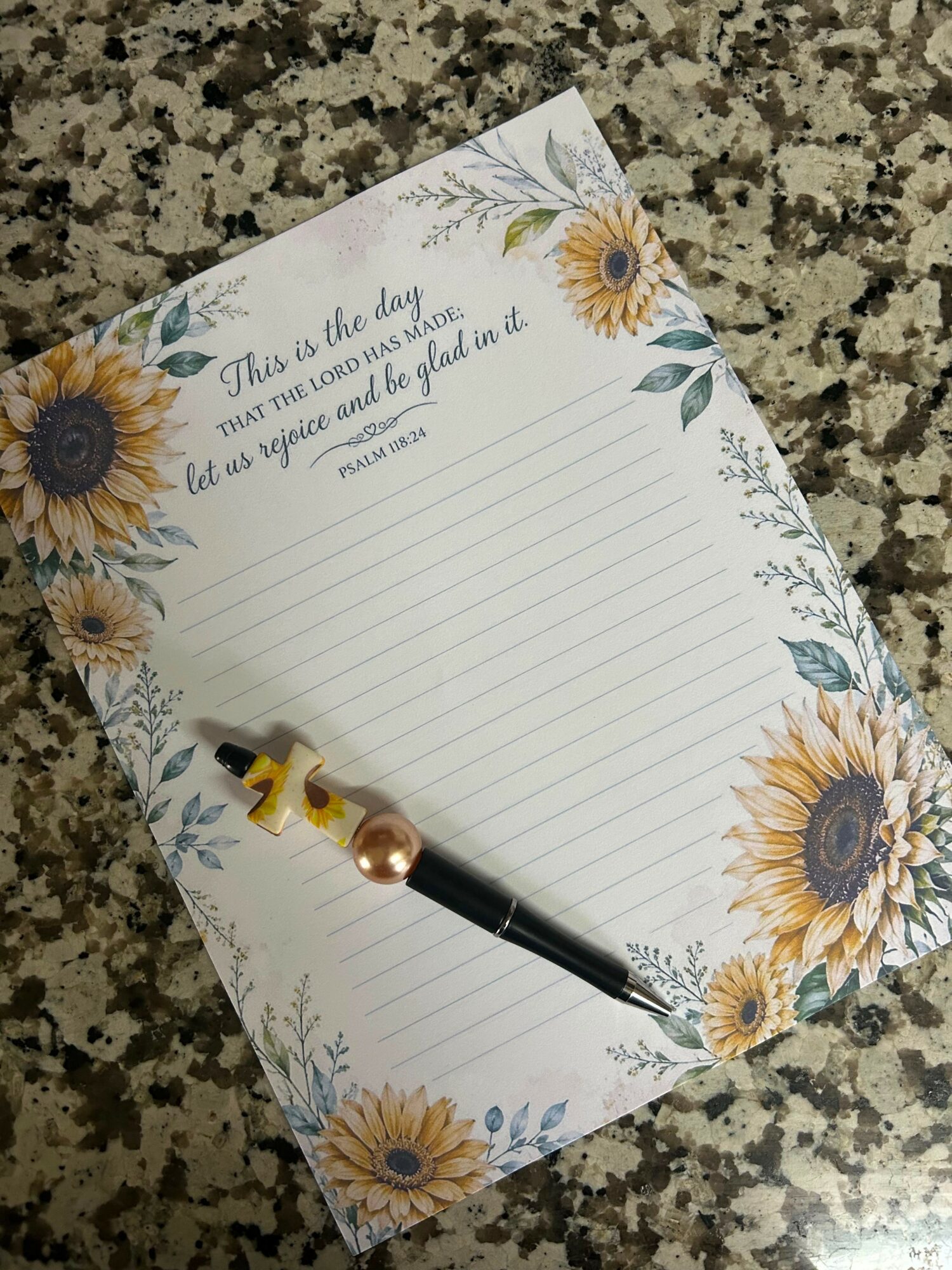 Notepad with sunflower design and a pen on a granite surface, lined for writing, with a quote at the top.