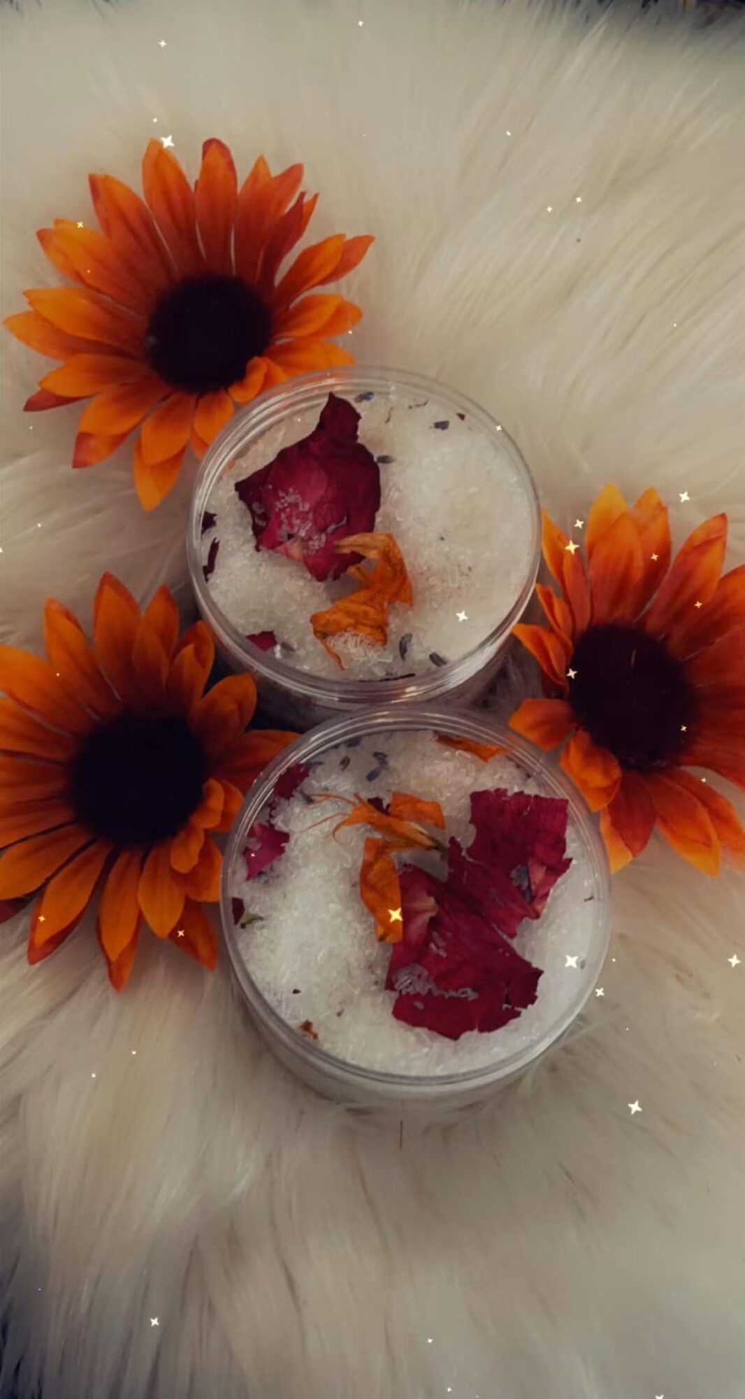 Two glasses with ice and red flower petals, surrounded by orange flowers on a white furry surface.