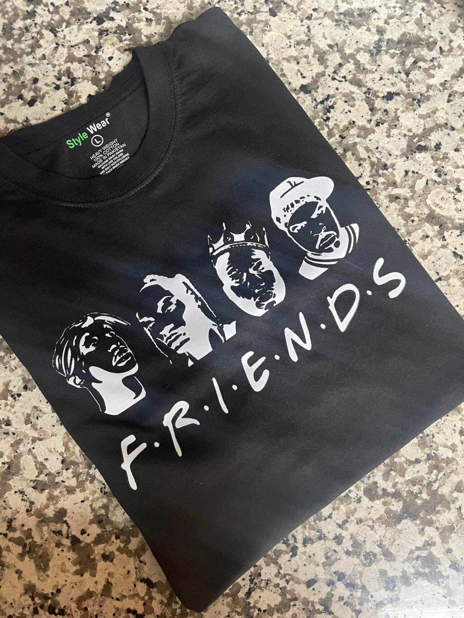 Black T-shirt with white graphic of four faces and the word 'F.R.I.E.N.D.S' below, laid on a speckled surface.