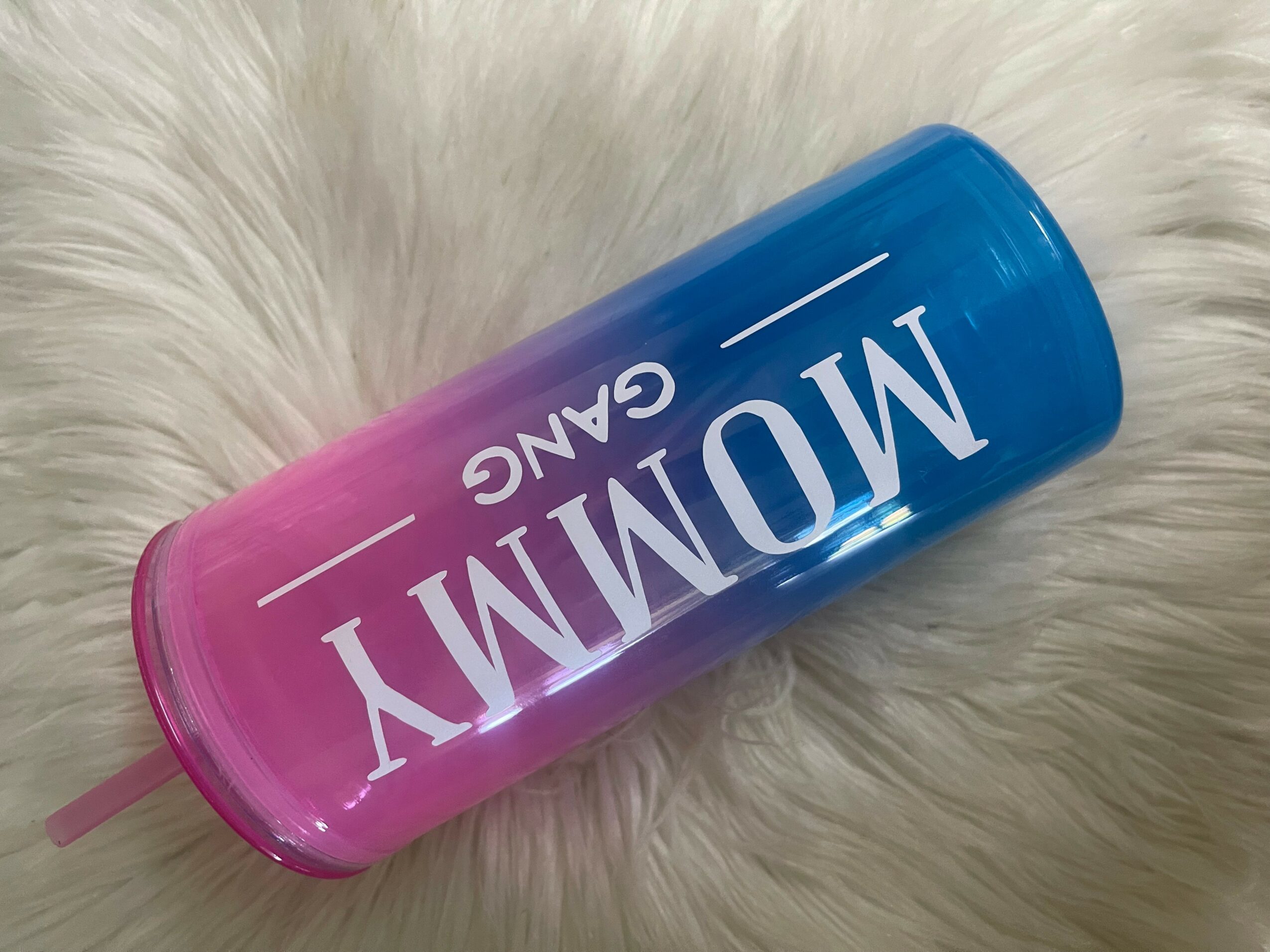 Colorful tumbler with pink and blue sections, labeled 'MOMMY GANG', lying on a white furry surface.