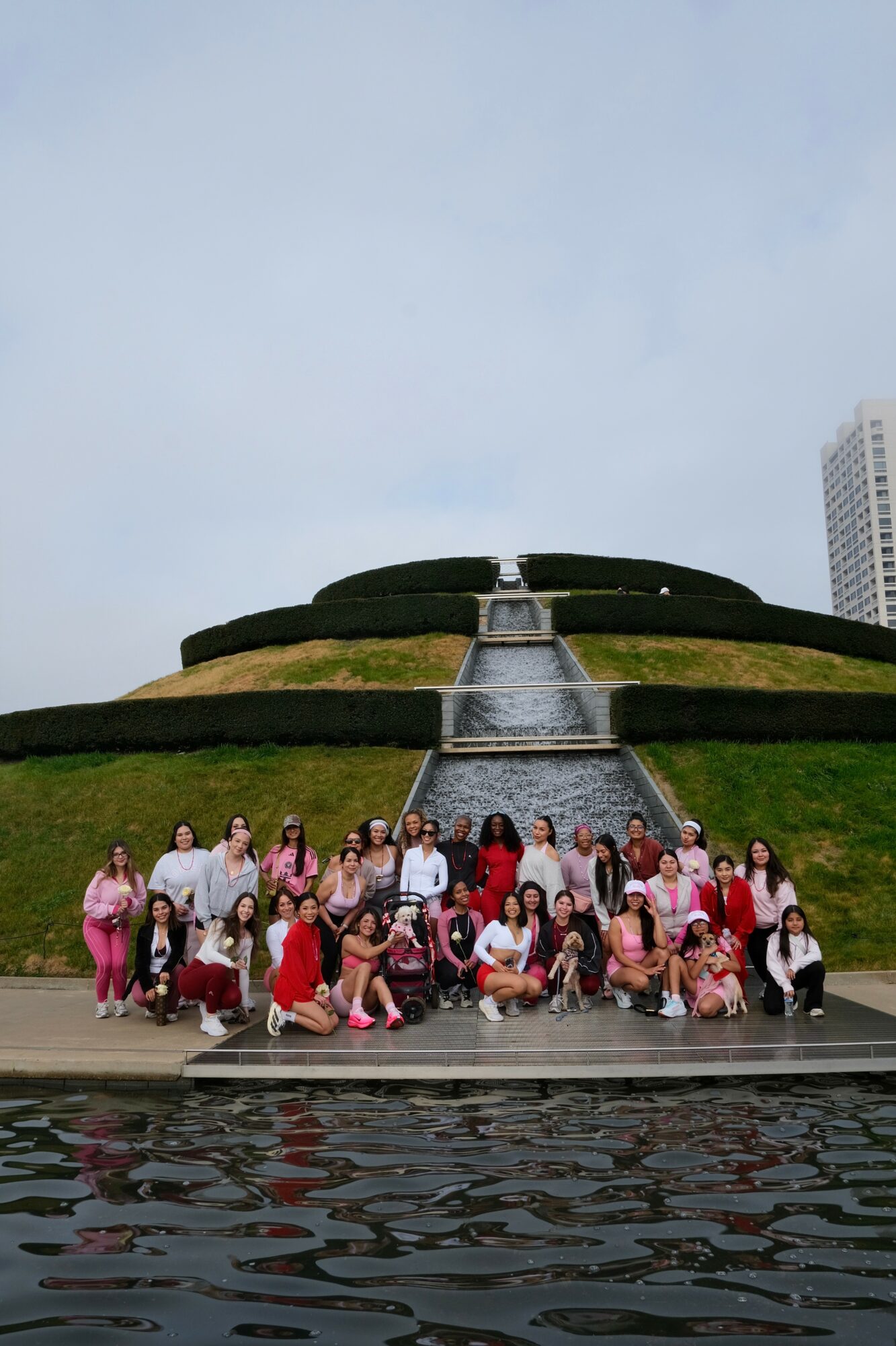 Group of people posing in front of a tiered landscaped fountain with a waterfall, water at the base, and tall buildings in the background.