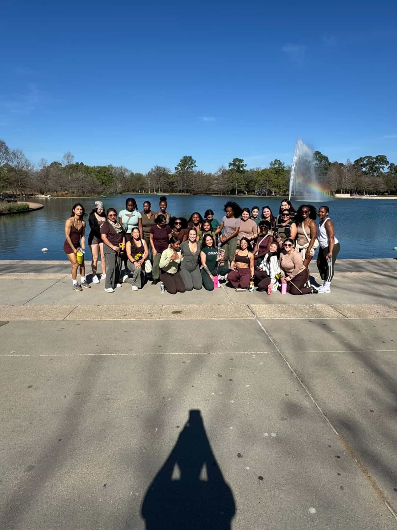 Group of people posing outdoors near a lake with a fountain, trees, and a clear blue sky in the background.