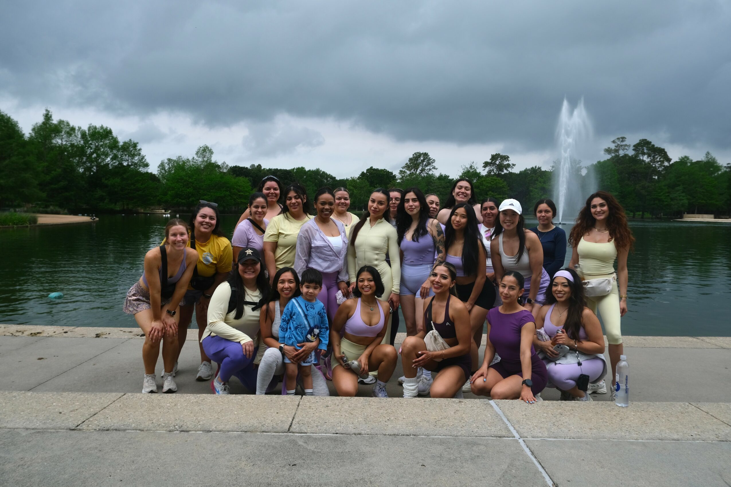 Group of women and children posing outdoors near a fountain with cloudy sky and trees in background.