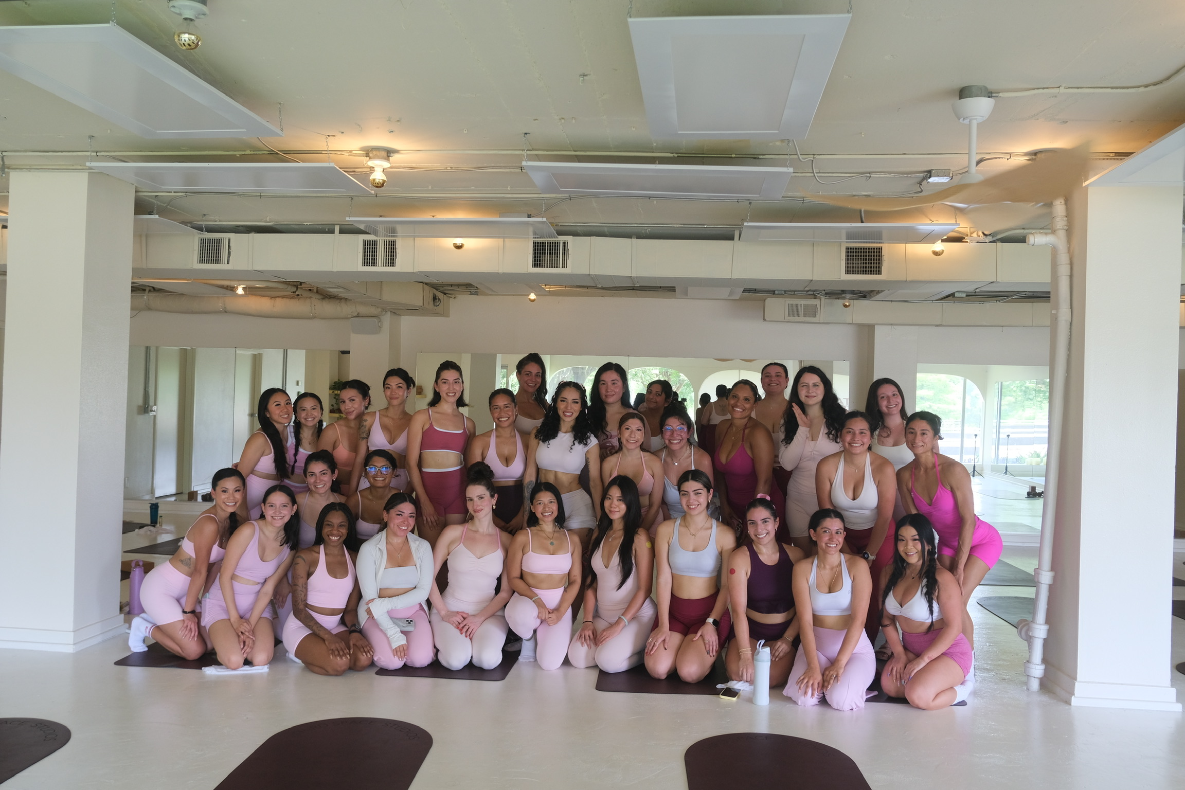 Group of women in dance costumes posing indoors, some kneeling and some standing, with a bright room background.