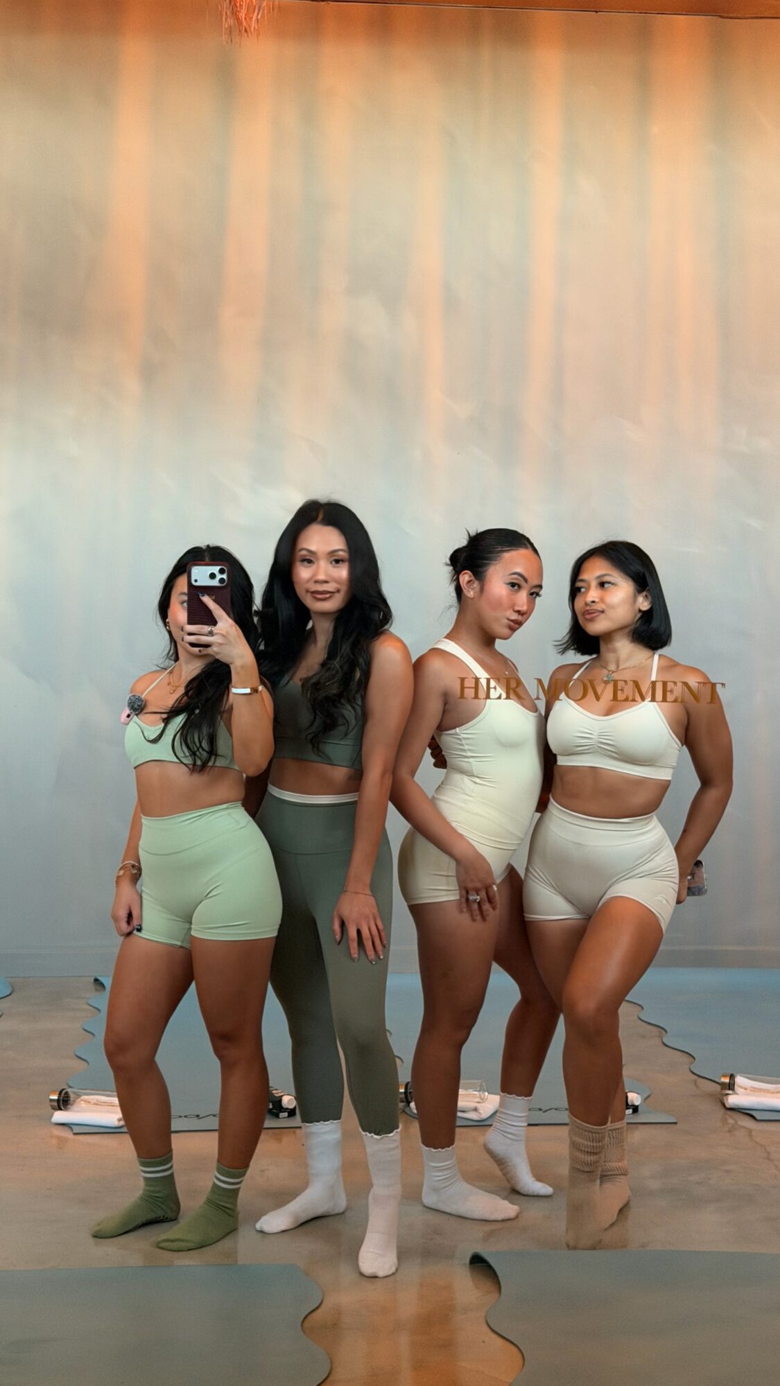 Four women in matching beige and green outfits pose in front of a colorful backdrop, with one taking a selfie.