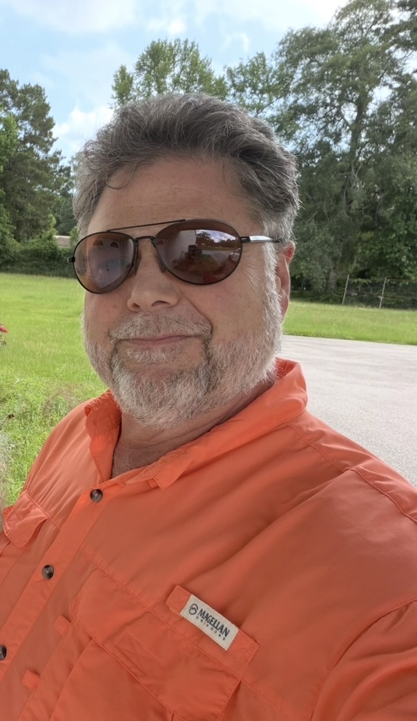 Man with sunglasses and gray hair outdoors, wearing an orange shirt, smiling, with trees and sky in background.