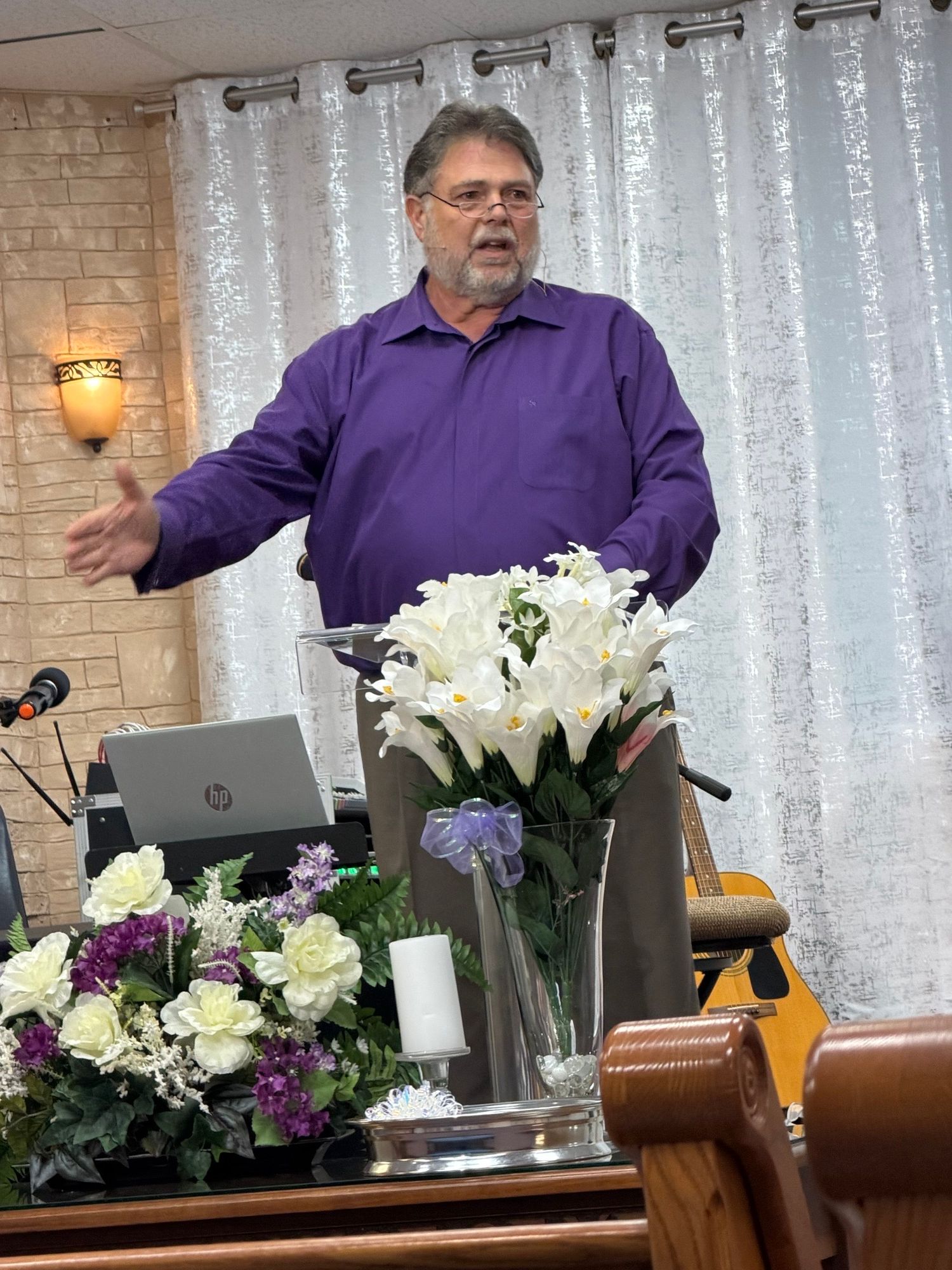 Man in purple shirt standing behind a table with a flower arrangement, laptop, and microphone, in a room with curtains.