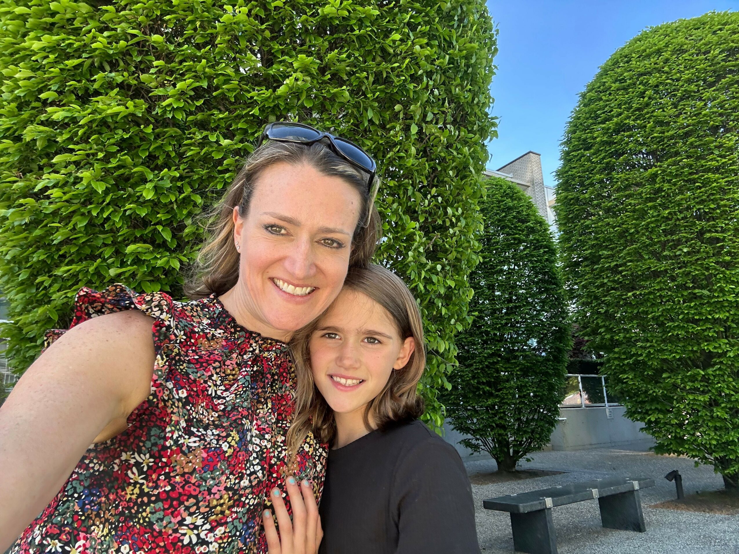 Two women lying on green bushes, smiling, with trees and a building in the background.