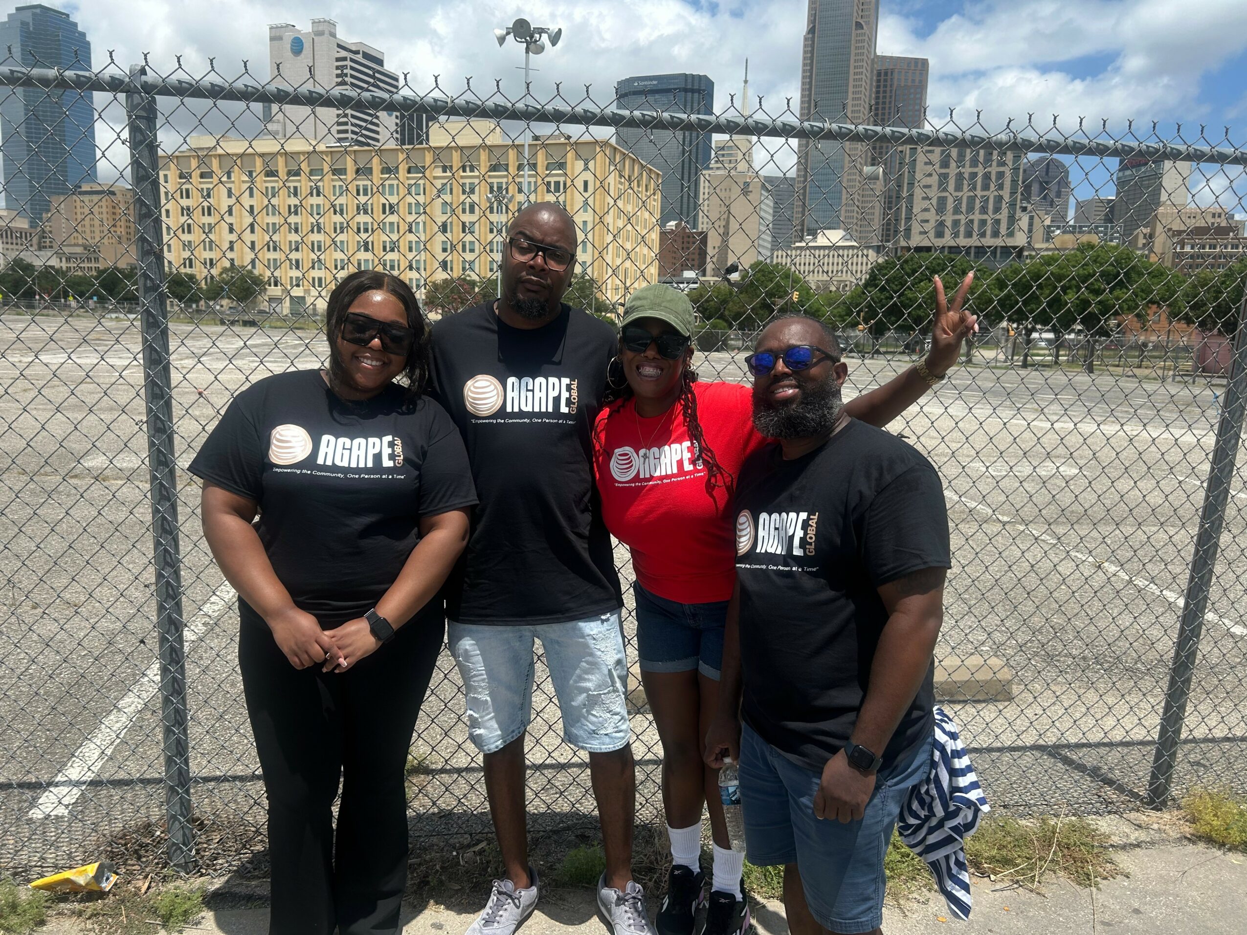 Four people standing outdoors in front of a chain-link fence with city buildings in the background, smiling and posing.