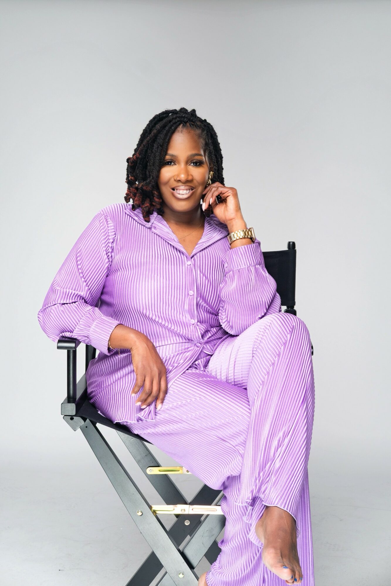 Woman with curly hair sitting on a chair, smiling, wearing a purple striped outfit, against a plain background.