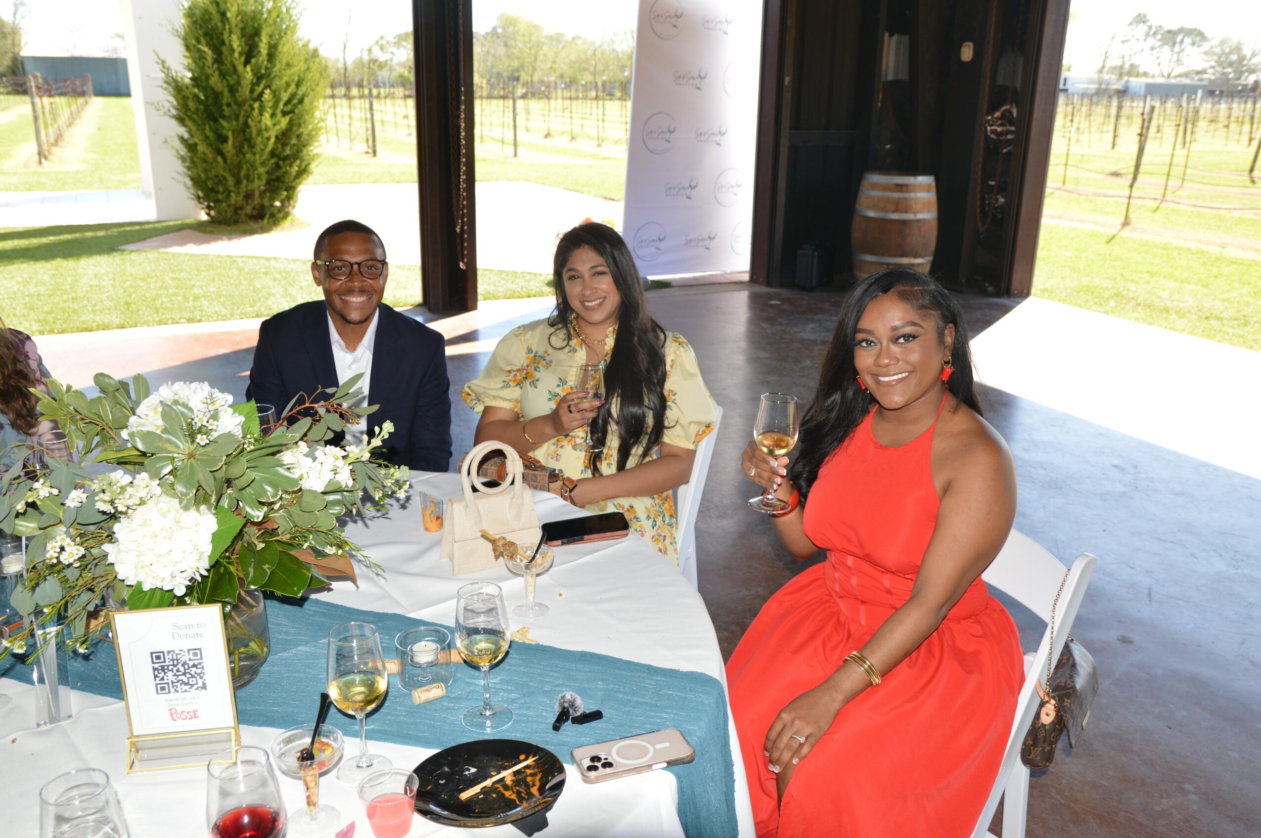 Three women seated at a table with flowers, drinks, and a purse, indoors with large windows showing outdoor scenery.