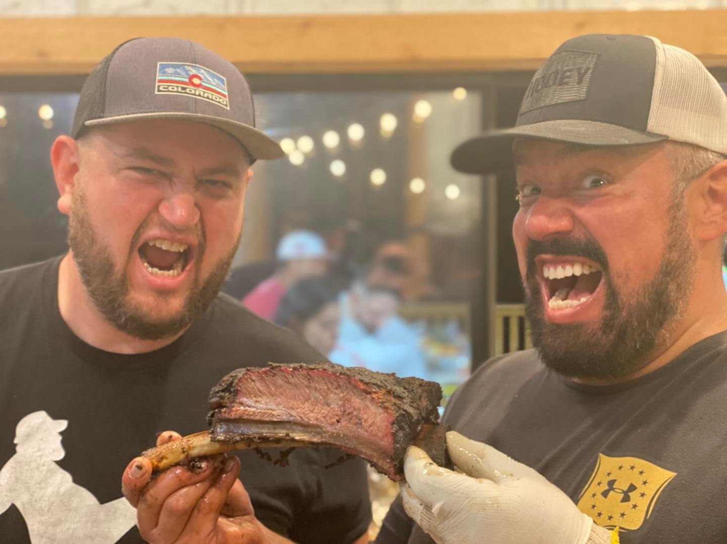Two men with beards and caps smiling and holding a large piece of cooked meat in a restaurant setting.