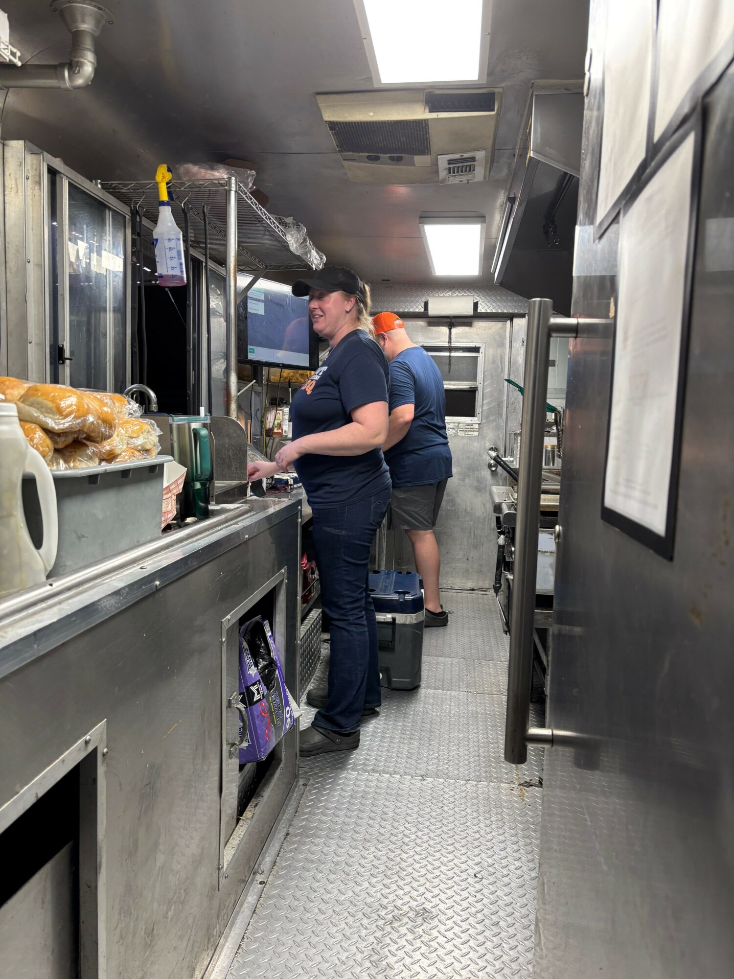 Two people in a commercial kitchen, one using a laptop, with stainless steel counters and equipment, and a window in the background.