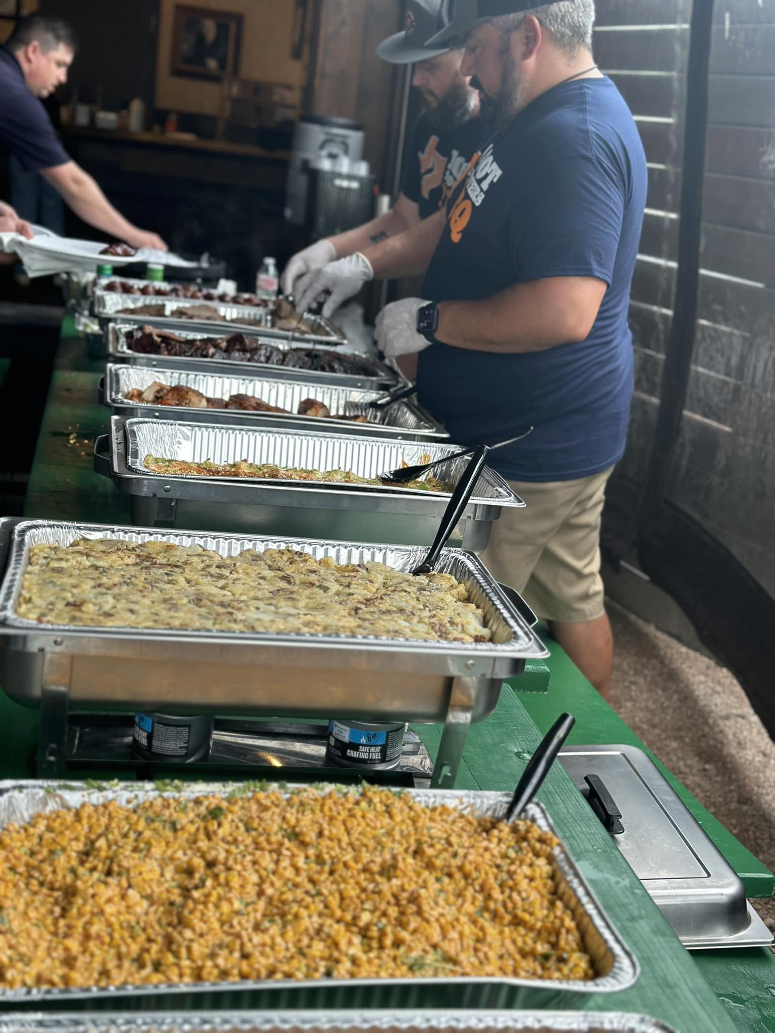 People serving food at a buffet with trays of various dishes on a green table, in a casual indoor setting.
