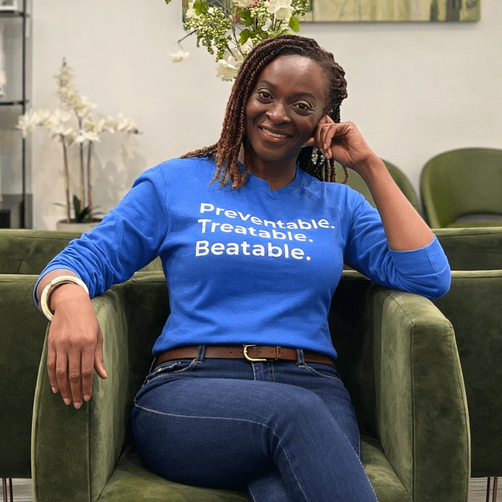 Woman sitting on green armchair, smiling, wearing a blue shirt with white text, in a room with plants and decor.