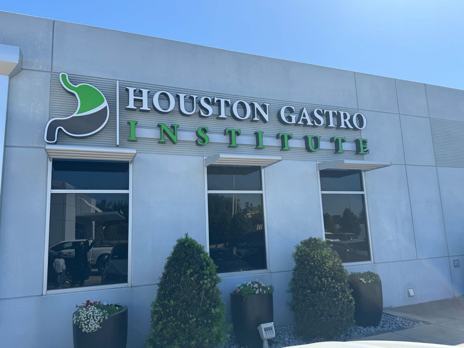 Exterior of Houston Gastro Institute building with sign and three windows, two bushes, and potted plants outside.